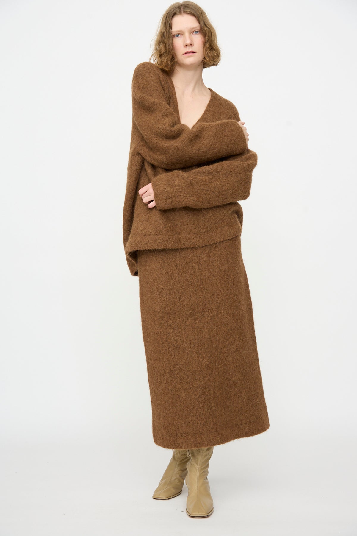 A person wearing the Lauren Manoogian Brushed V Neck in Rust, paired with a matching skirt and beige boots, stands against a plain white background.
