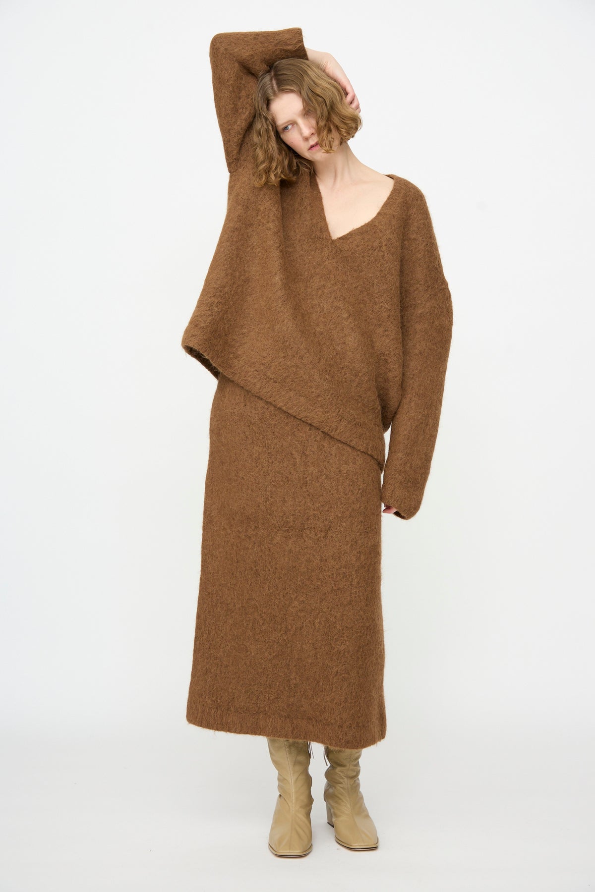 A person wearing the Lauren Manoogian Brushed V Neck in Rust and a matching skirt with beige boots stands against a plain white background, posing with one arm raised.
