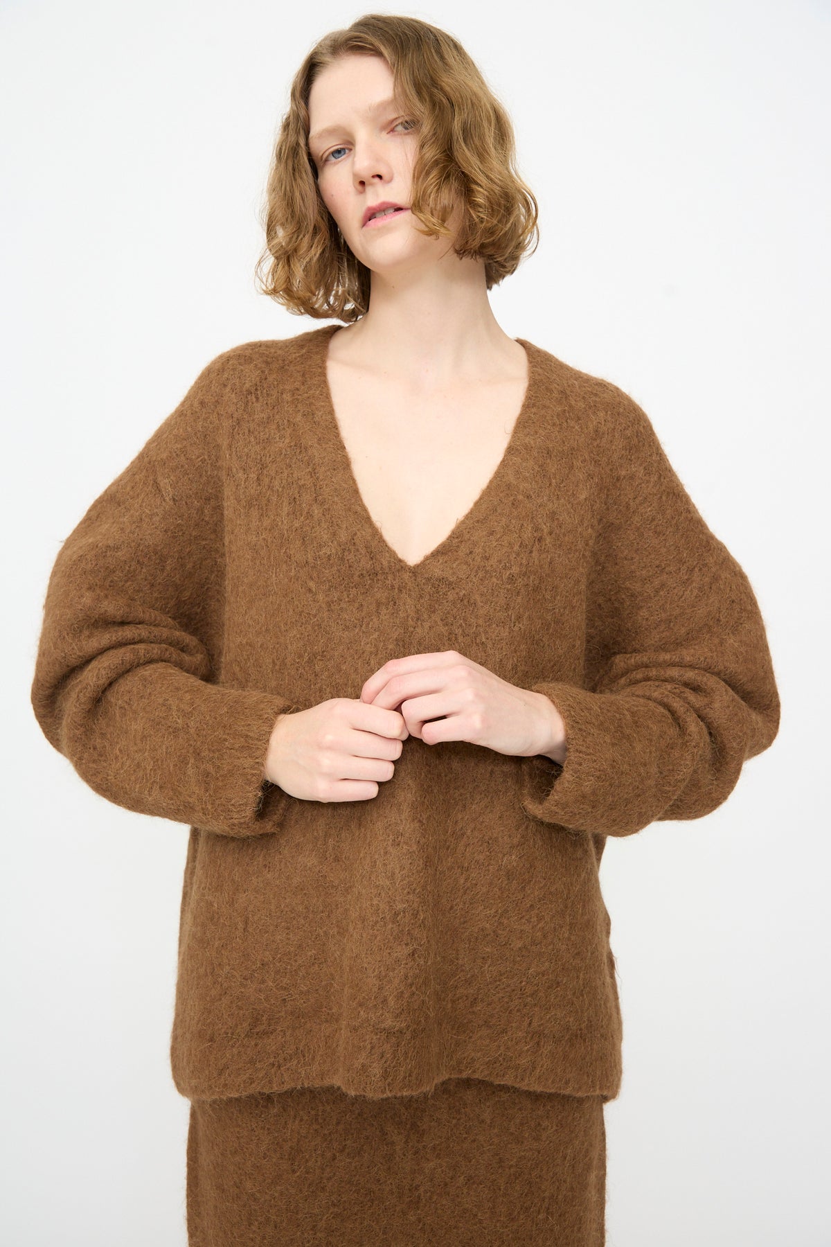 A person with short, wavy blond hair is wearing an oversized-fit Lauren Manoogian Brushed V Neck sweater in Rust, paired with a matching skirt, and standing against a plain white background.