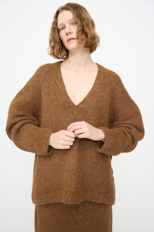 A person with short, wavy blond hair is wearing an oversized-fit Lauren Manoogian Brushed V Neck sweater in Rust, paired with a matching skirt, and standing against a plain white background.
