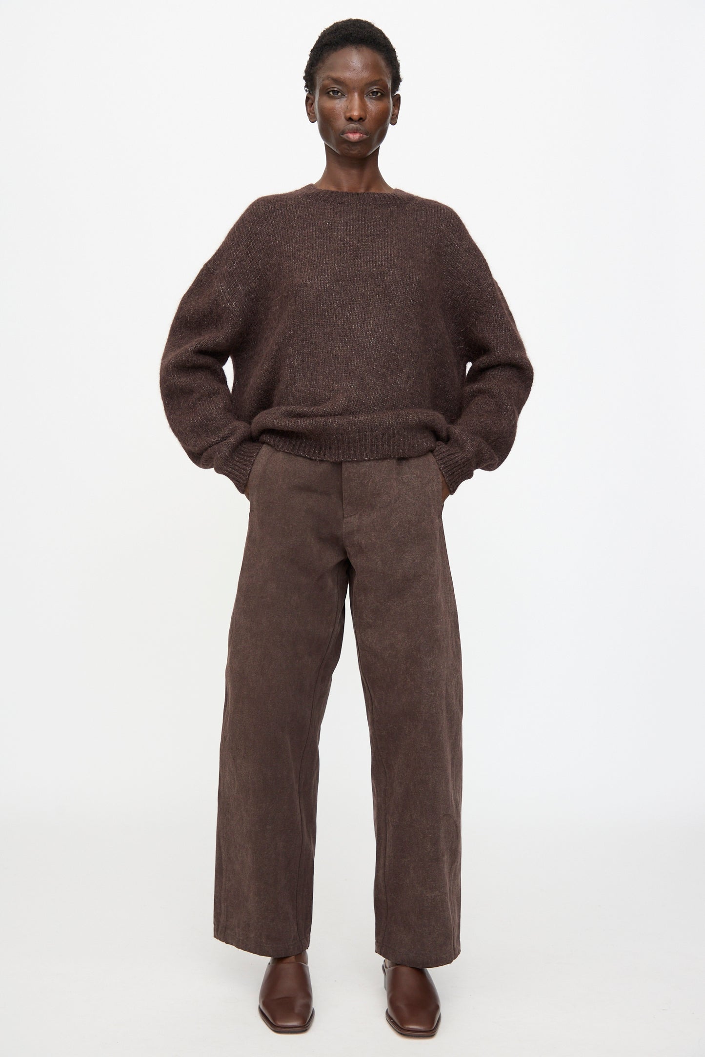 A person stands against a white background wearing a loose brown sweater, Lauren Manoogian Canvas Pants in Washed Soil (hand-dyed cotton, high waist, curved leg), and brown shoes, with hands in pockets.