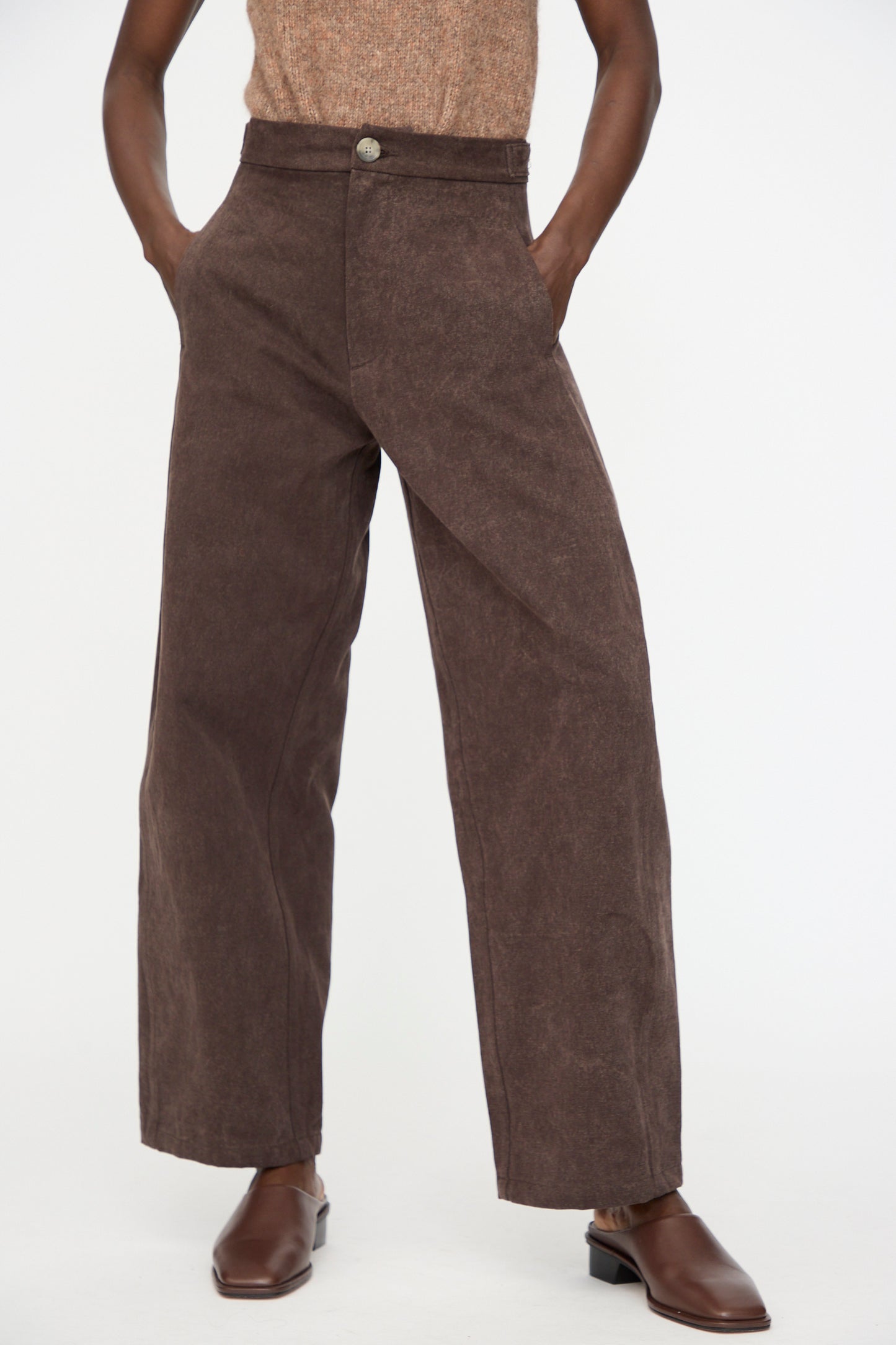 A person wearing Lauren Manoogian's Canvas Pants in Washed Soil—a pair of high-waist, wide-leg brown pants—stands with hands in pockets and brown shoes against a plain background.