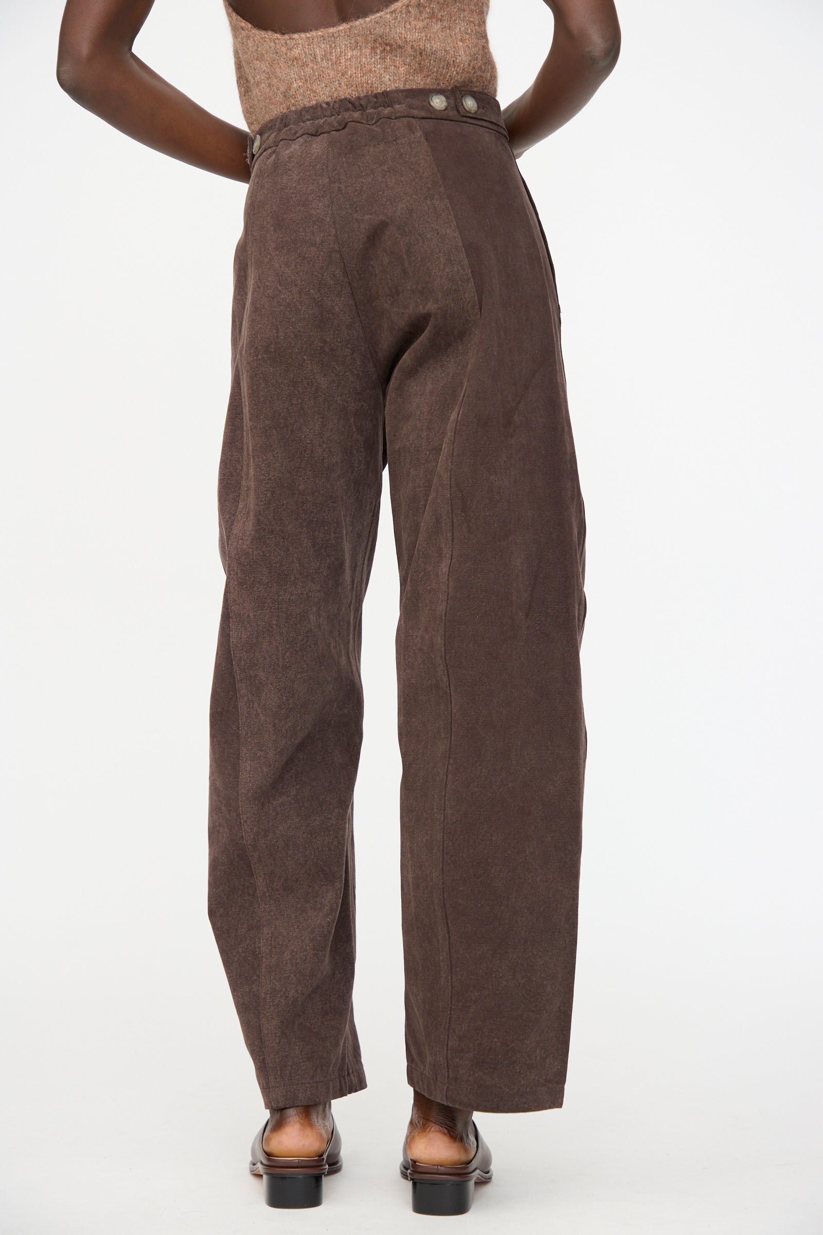 Someone stands with their back to the camera against a plain background, wearing Lauren Manoogian Canvas Pants in Washed Soil—high-waisted, curved leg pants with side buttons, crafted from hand-dyed cotton.