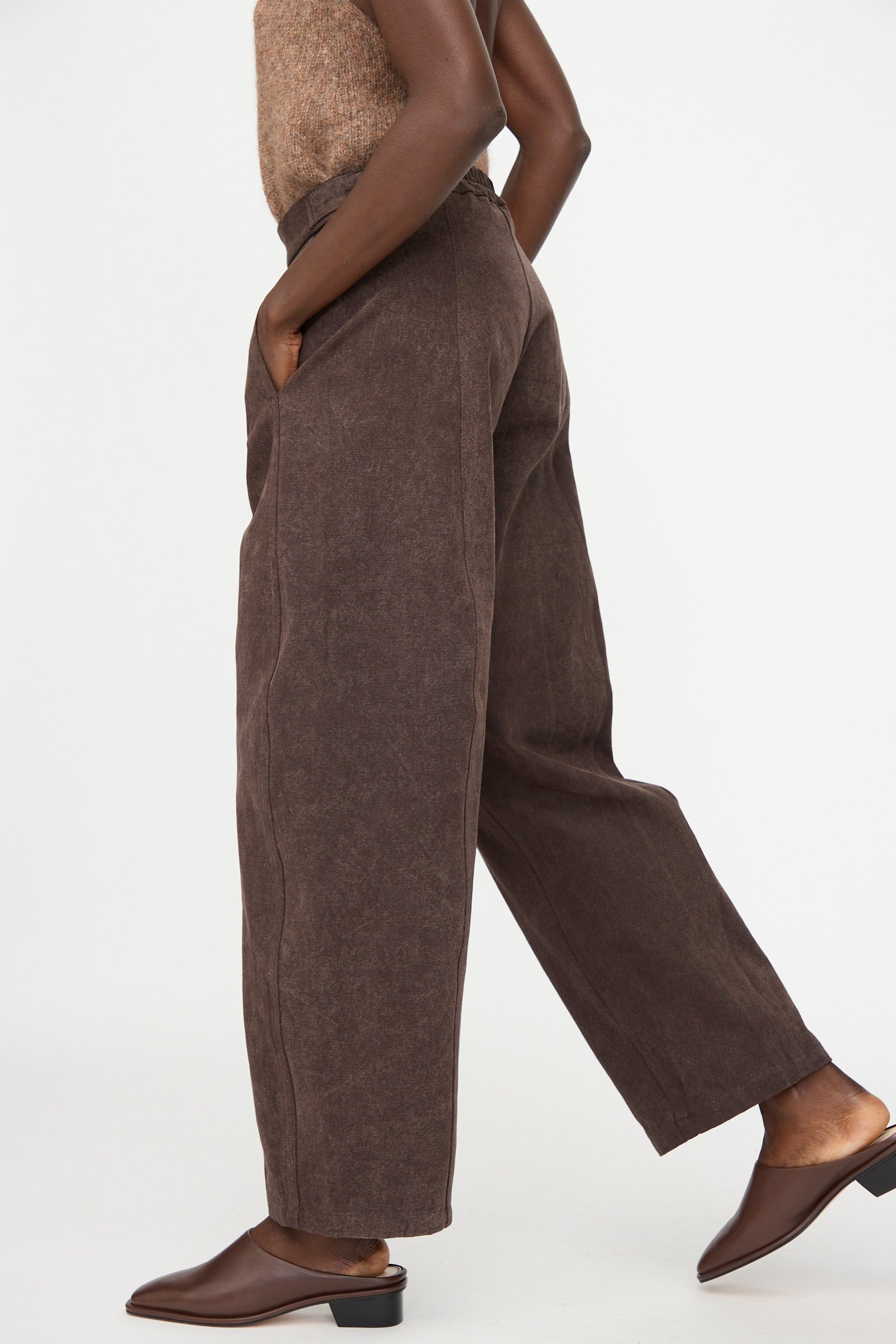 A person wears Lauren Manoogian’s Canvas Pants in Washed Soil, paired with a brown sleeveless top and brown heeled mules, standing with one foot forward against a plain white background.
