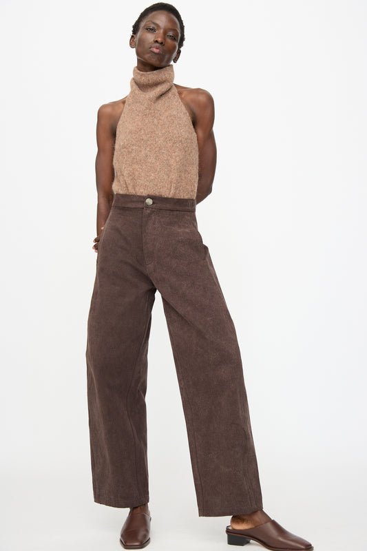 A person stands against a plain white background wearing a sleeveless, high-neck brown sweater, Lauren Manoogian Canvas Pants in Washed Soil, and brown shoes.
