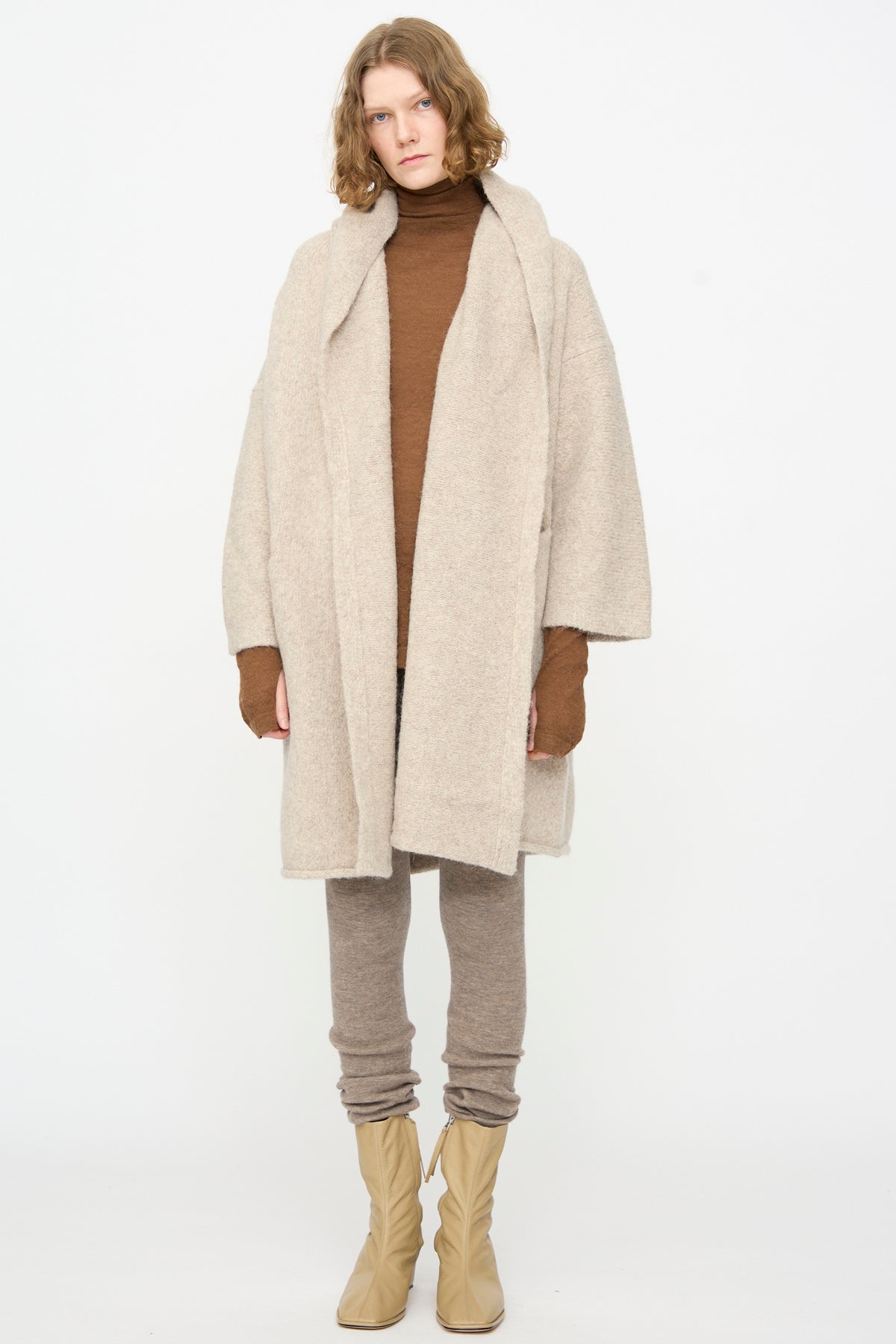 A person stands against a white background, wearing the Lauren Manoogian Capote Coat in Pebble—an oversized, hand-loomed alpaca wool coat made in Peru—paired with a brown turtleneck, taupe leggings, and tan boots.