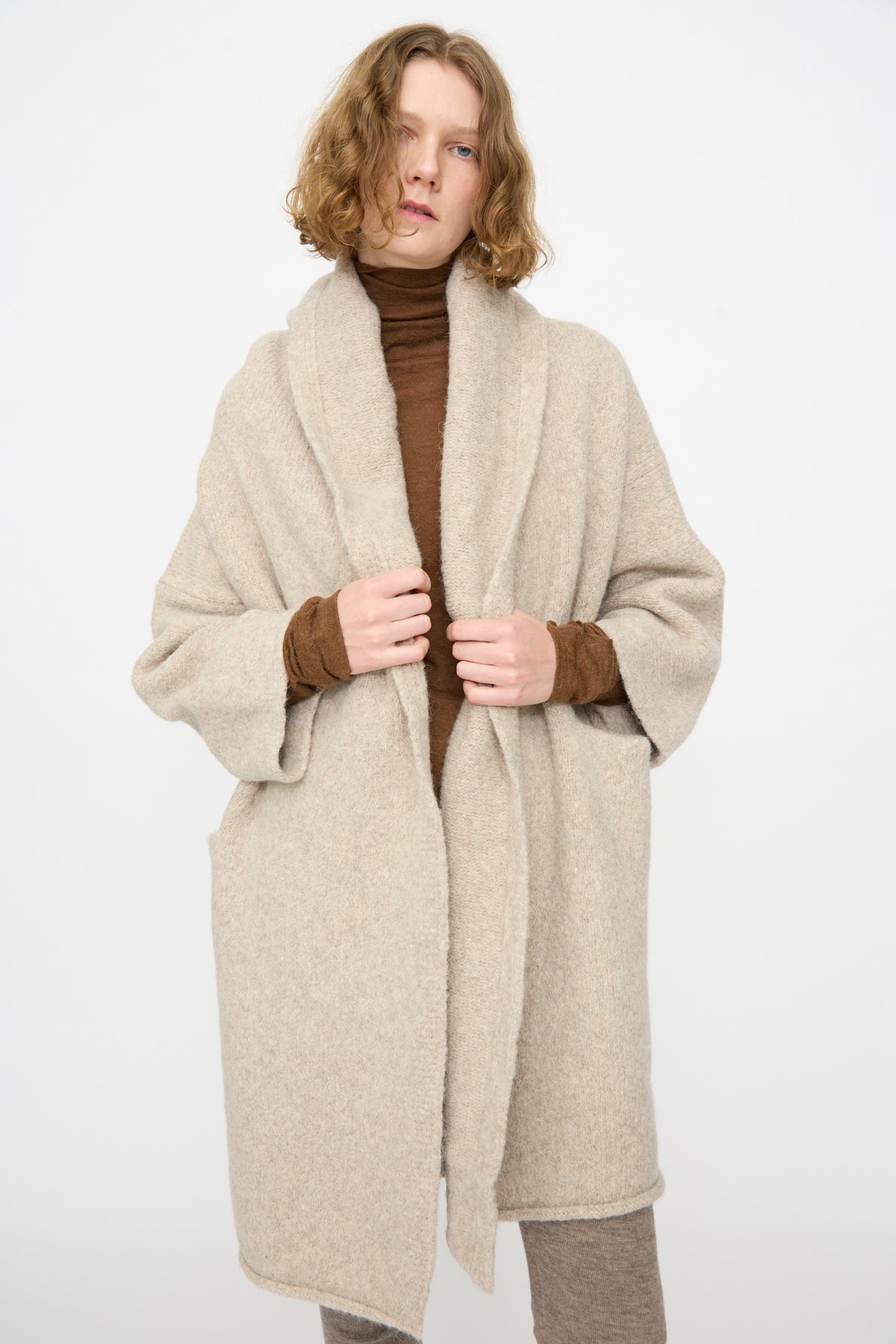A person wearing the Lauren Manoogian Capote Coat in Pebble—a beige, oversized, hand-loomed piece—layered over a brown turtleneck and beige pants, stands against a plain white background. The look evokes effortless style reminiscent of Peruvian-made alpaca wool garments.