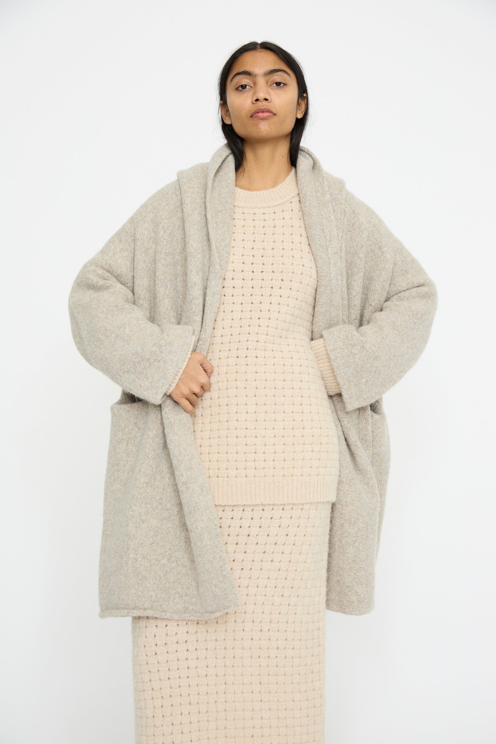 A model wearing the Lauren Manoogian Capote Coat in Pebble, styled with a beige knitted dress, stands against a plain white background, facing forward. 