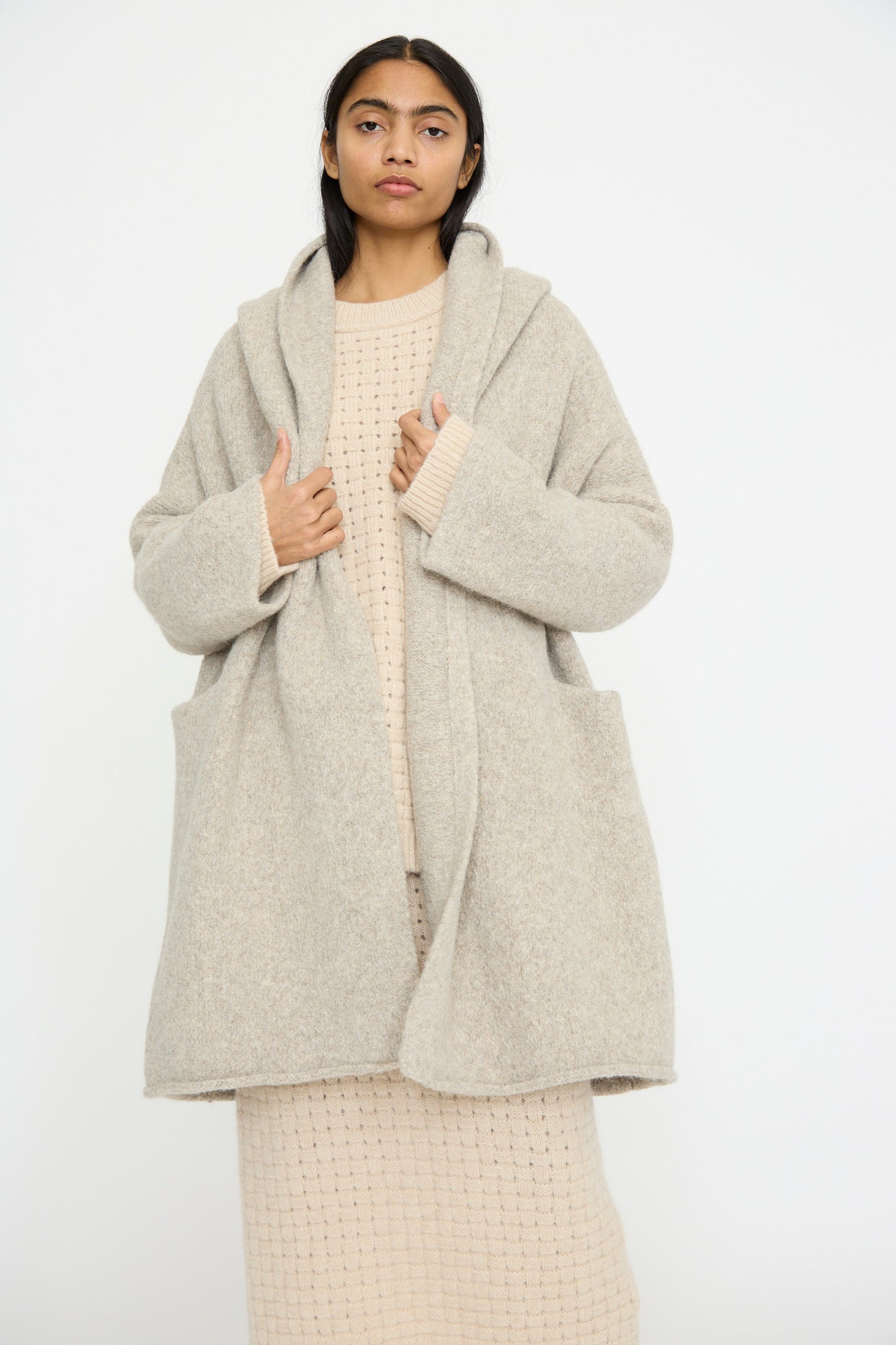 A model displays the Lauren Manoogian Capote Coat in Pebble, a relaxed fit long beige coat made from alpaca wool, over a matching knit dress against a plain white background.