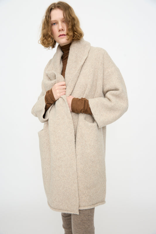 A person with shoulder-length wavy hair is wearing the Lauren Manoogian Capote Coat in Pebble, a hand-loomed oversized alpaca wool coat made in Peru. The coat is styled with a brown turtleneck and matching pants, and the individual stands against a plain white background.