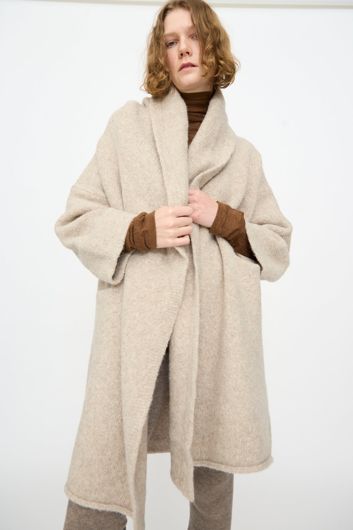 A person wearing the Lauren Manoogian Capote Coat in Pebble, styled over a brown turtleneck and beige pants, stands against a plain white background.