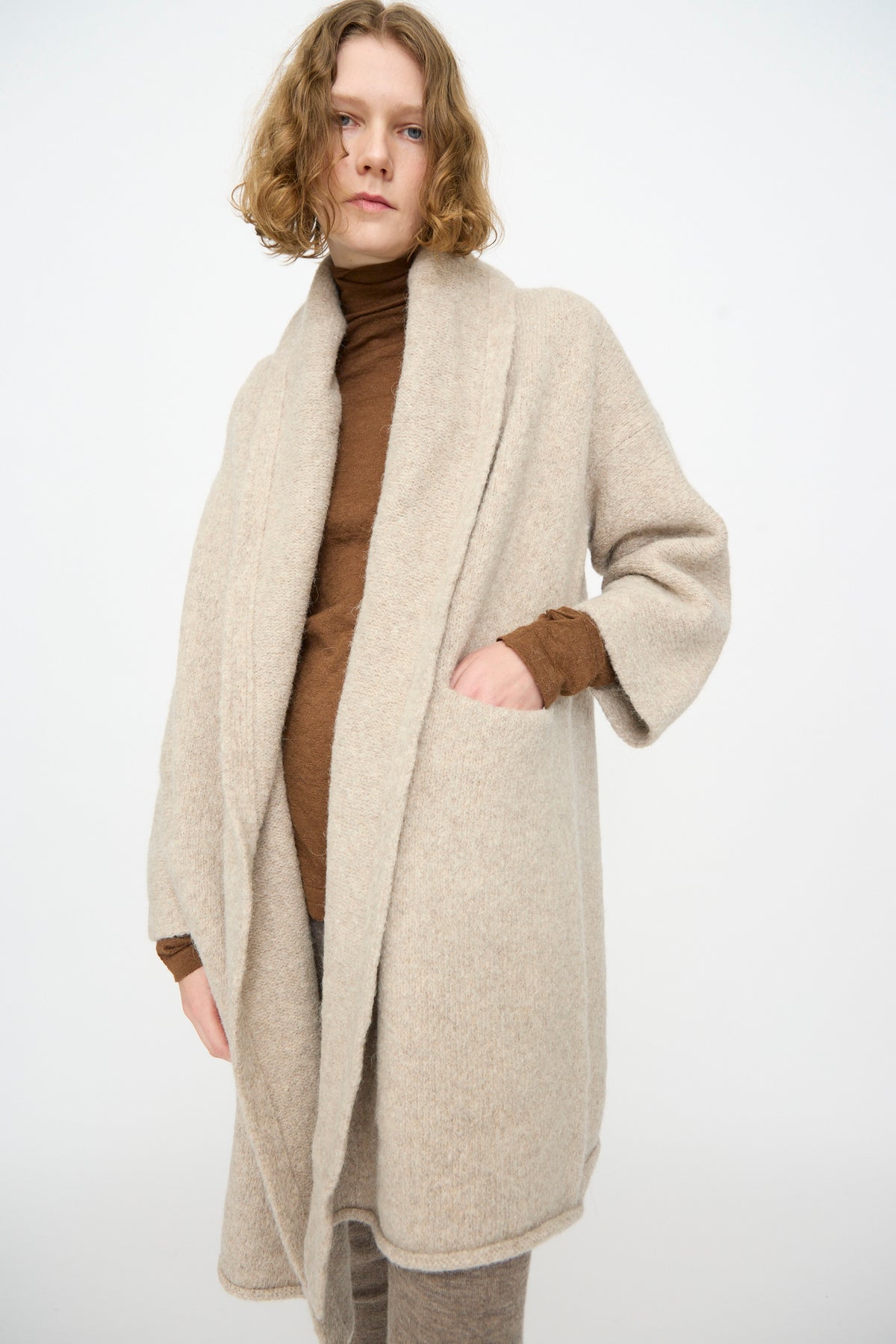 A person with shoulder-length curly hair wears the Lauren Manoogian Capote Coat in Pebble—a beige, hand-loomed open-front cardigan from Peru—layered over a brown turtleneck and beige pants, standing against a plain white background.