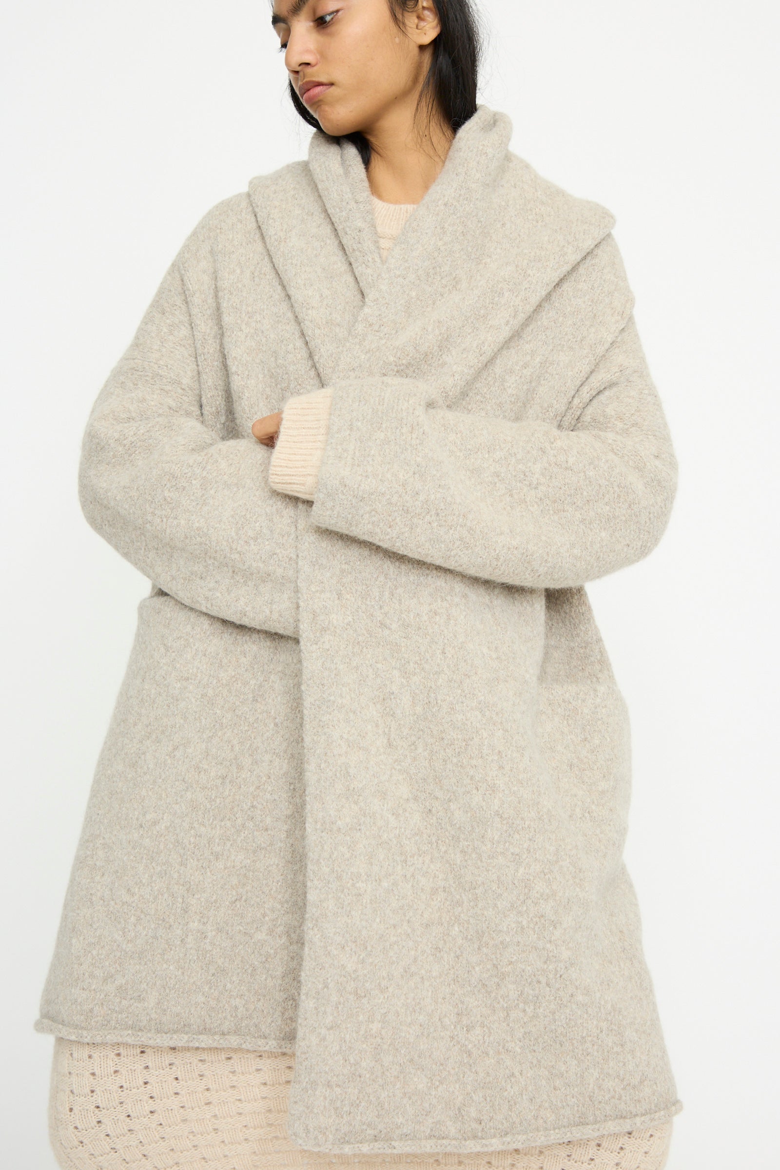 A person is wrapped in the cozy, oversized Lauren Manoogian Capote Coat in Pebble with a textured finish over a knitted sweater, against a plain background. 