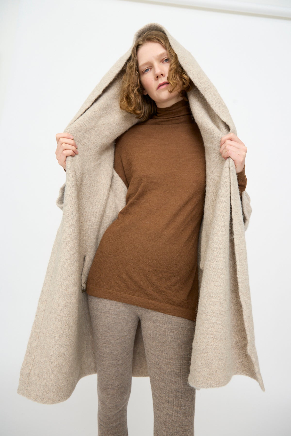 A person wearing a brown turtleneck and beige pants stands holding open the Lauren Manoogian Capote Coat in Pebble—a hand-loomed alpaca wool coat made in Peru—while looking directly at the camera against a plain light background.