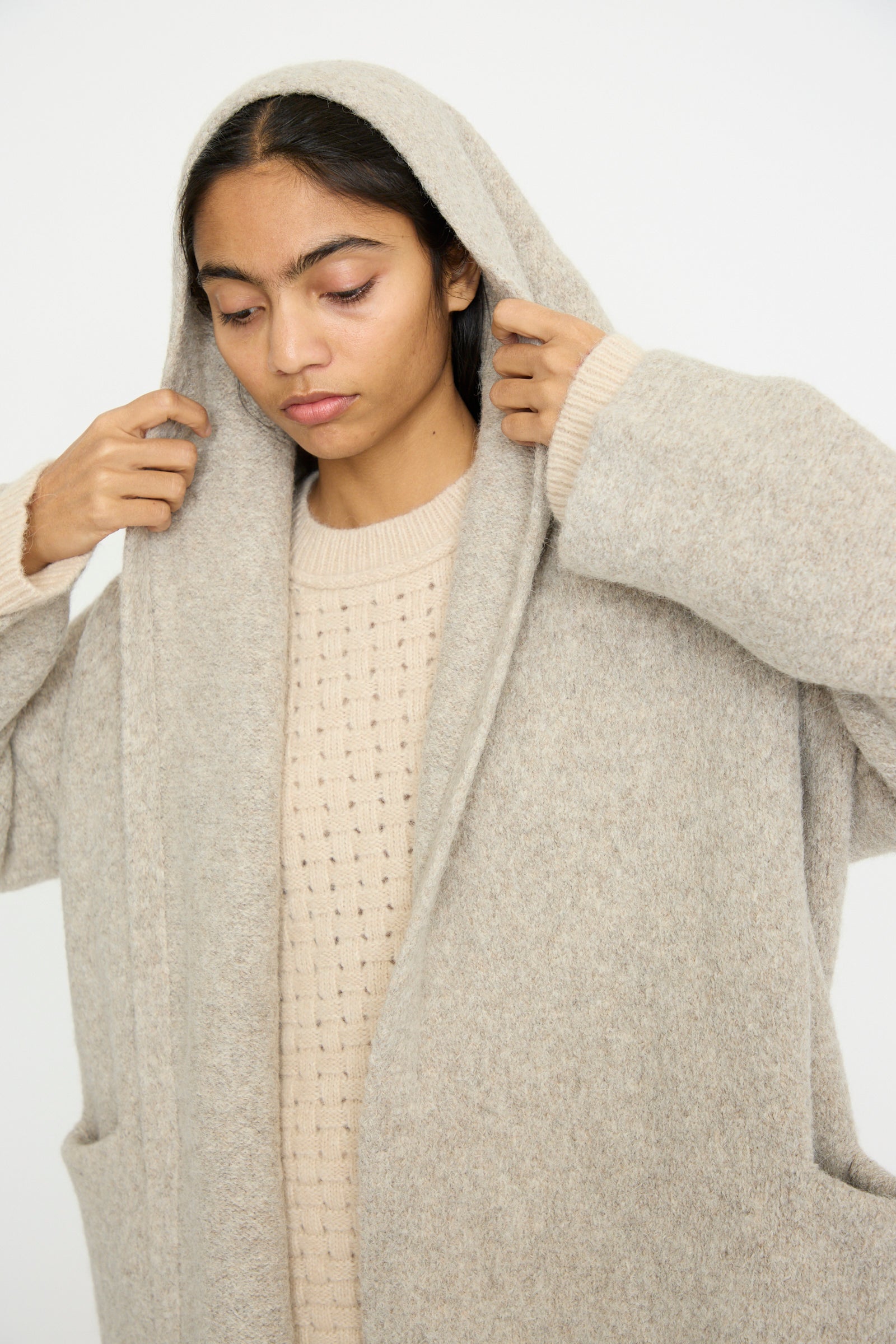 A person adjusts the hood of a Lauren Manoogian Capote Coat in Pebble, looking down and wearing neutral-toned clothing against a plain background.