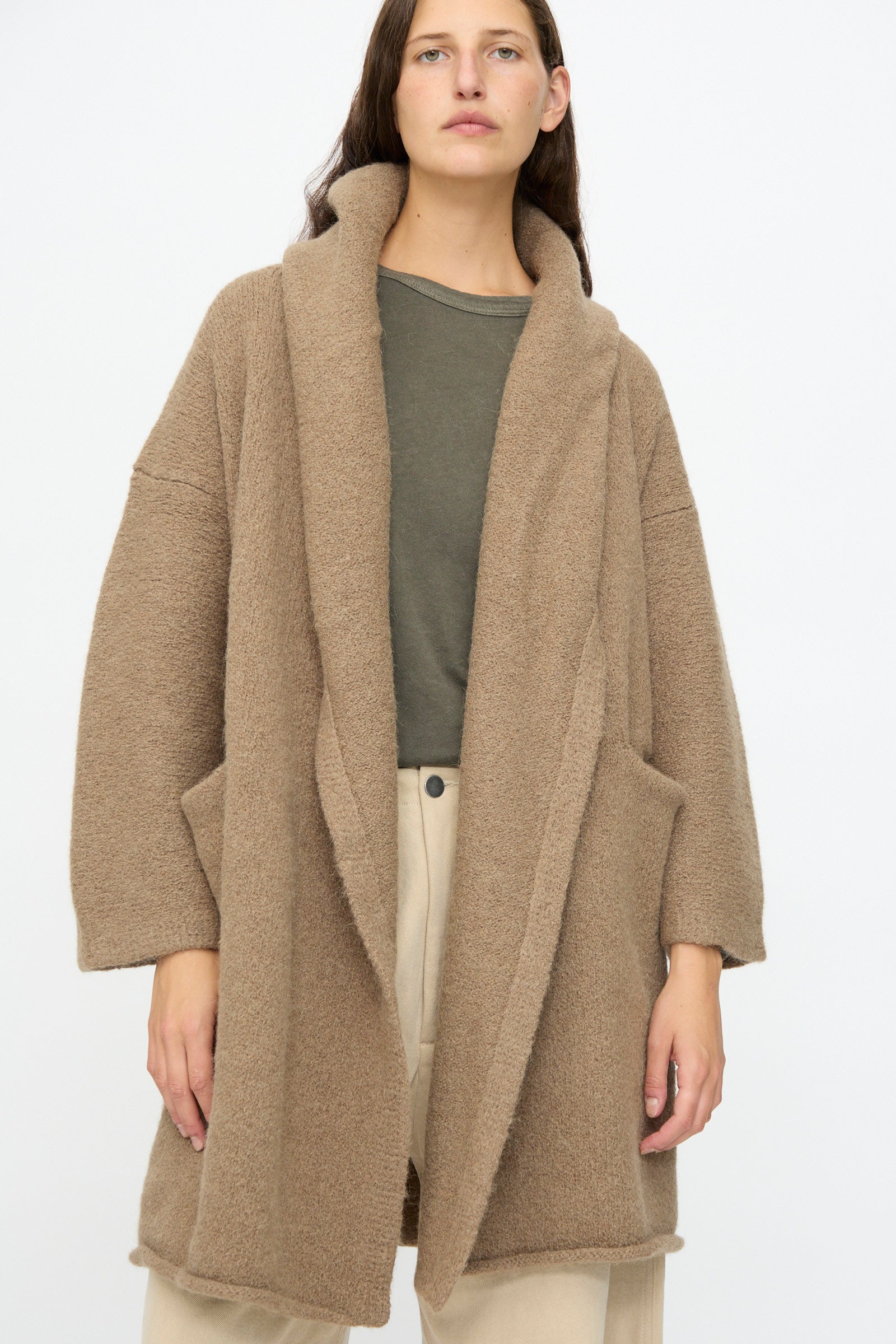 A person wears the Lauren Manoogian Capote Coat in Tobacco—a relaxed fit alpaca wool piece—over a green shirt and beige pants, standing against a plain light background.