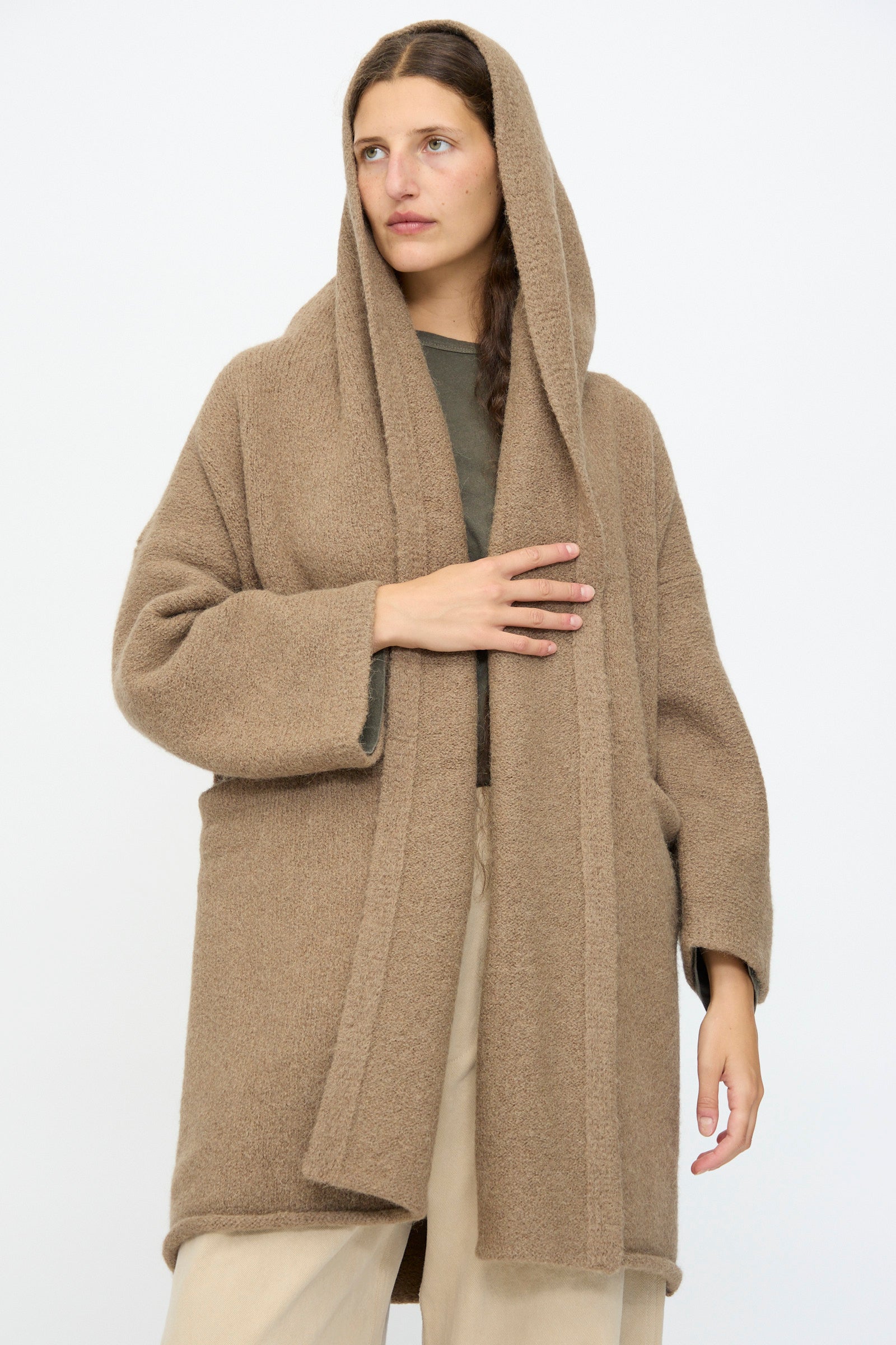 A woman wears the Lauren Manoogian Capote Coat in Tobacco—a relaxed fit brown hooded alpaca wool coat—over a green top and beige pants, standing against a plain white background.
