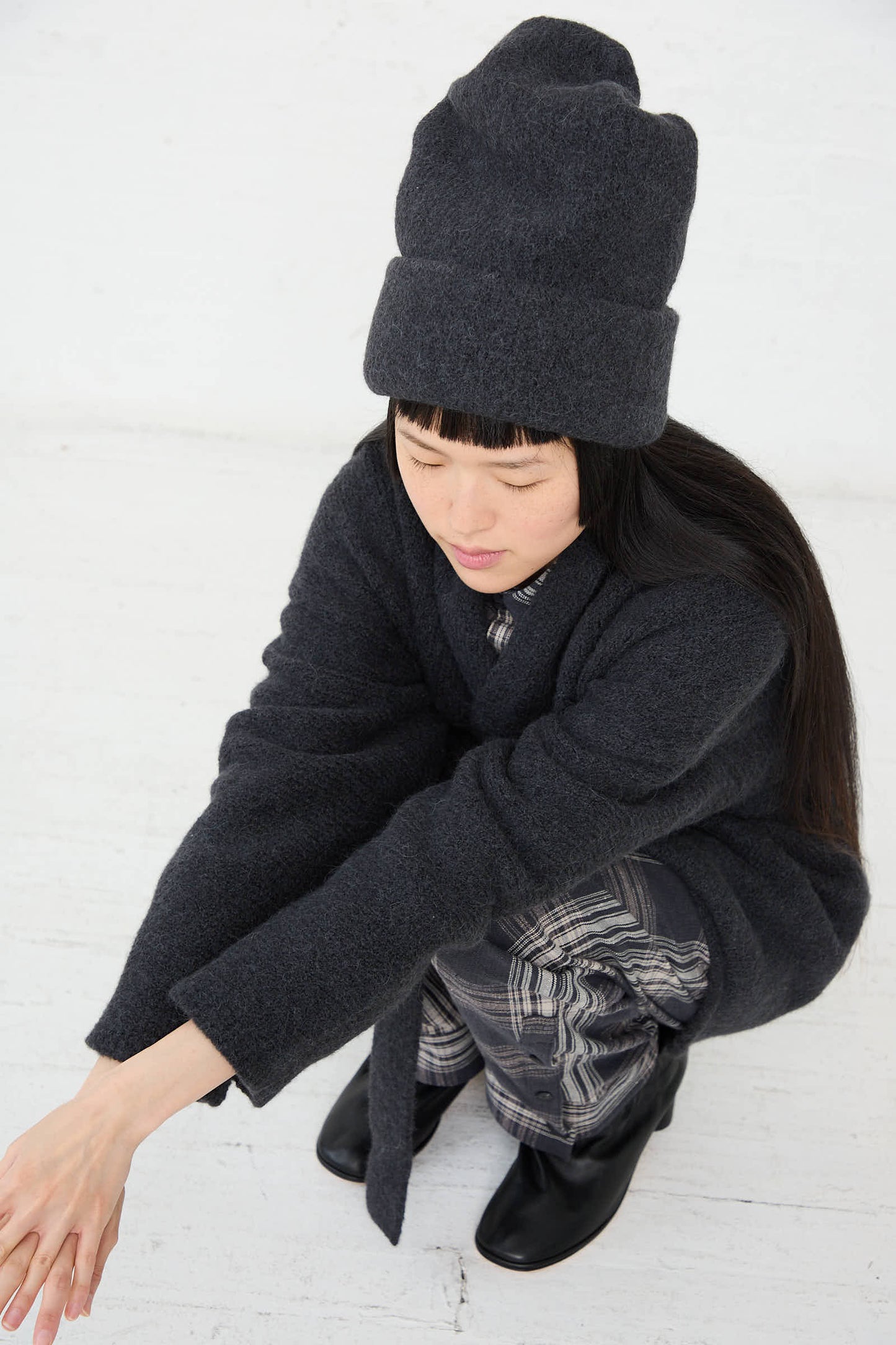 A person wearing a dark coat, the Carpenter Hat in Ink by Lauren Manoogian, and patterned pants squats on a white floor, looking downwards.