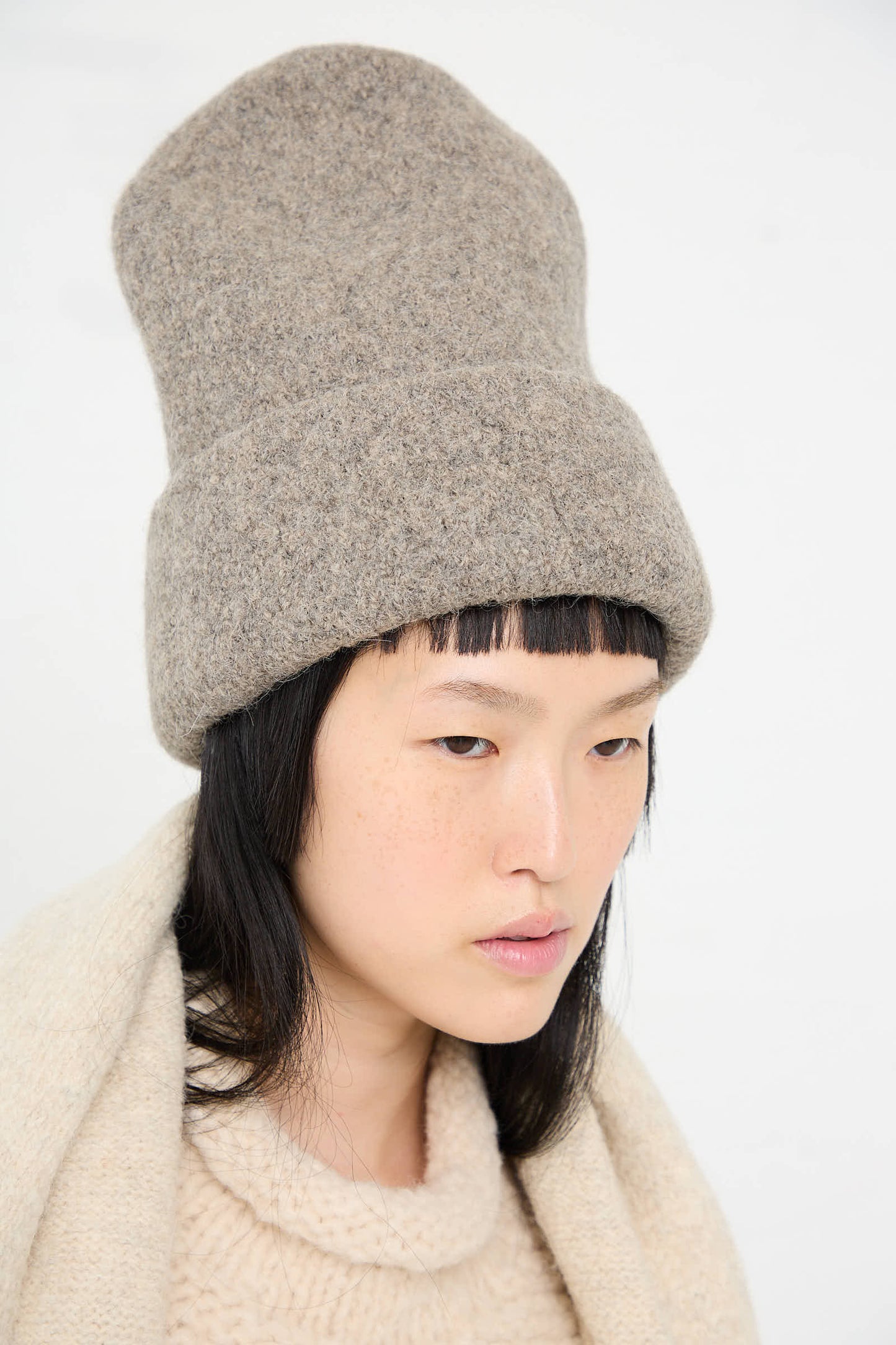 Person with straight black hair, wearing the Lauren Manoogian Carpenter Hat in Rock and a matching woolen sweater and scarf. The background is plain white.