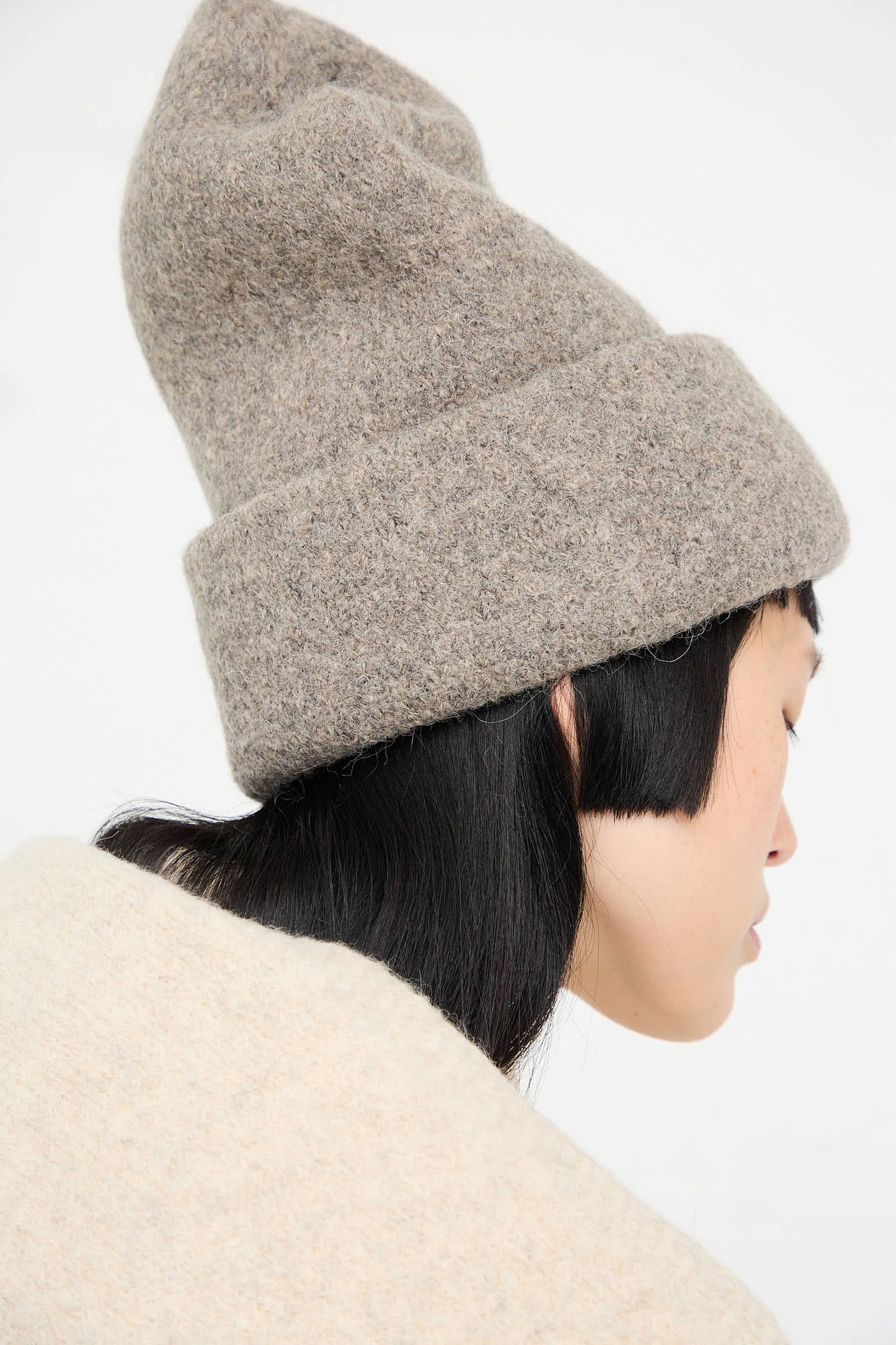 A person with long dark hair is wearing a light brown Carpenter Hat in Rock by Lauren Manoogian, an alpaca beanie made in Peru, and a beige garment, facing away from the camera.