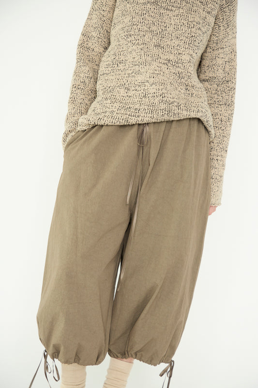 A person wears a textured beige sweater and Lauren Manoogian’s Cinch Pants in Mole, featuring wide legs and drawstring cuffs, made from 100% cotton, against a plain background.