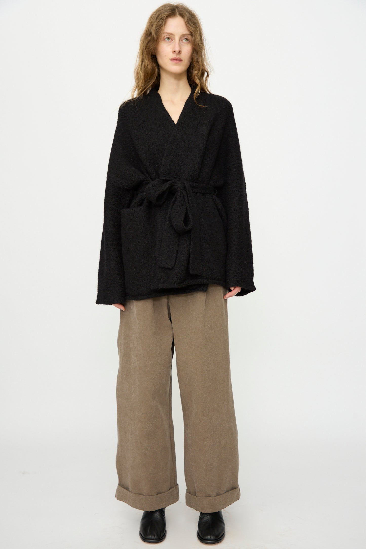 A person stands facing forward against a plain white background wearing the Lauren Manoogian Coto Cardigan in Black, paired with wide-leg beige cuffed pants and black shoes.