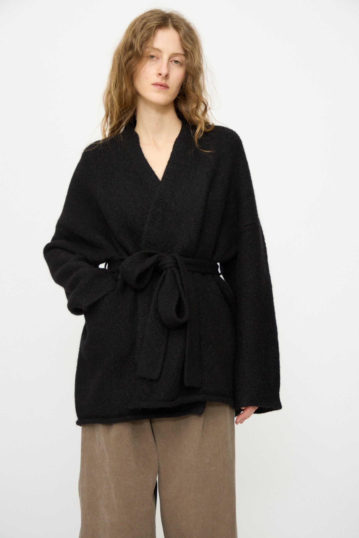 A person wears the Lauren Manoogian Coto Cardigan in Black, loosely belted at the waist, paired with wide-leg taupe pants, standing against a plain white background.