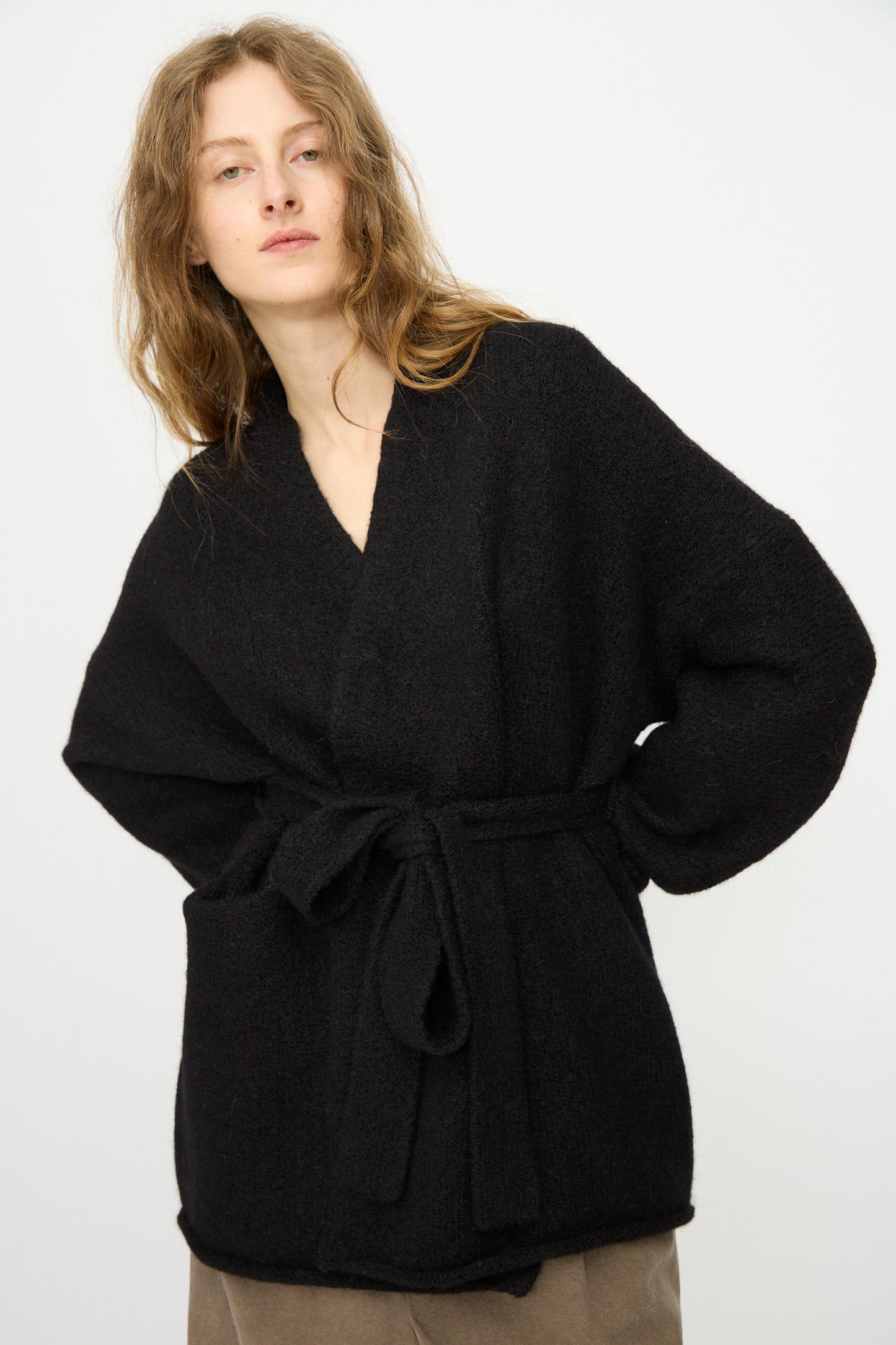A person with long, wavy hair wears the Lauren Manoogian Coto Cardigan in Black, featuring large front pockets, and stands against a plain light background.