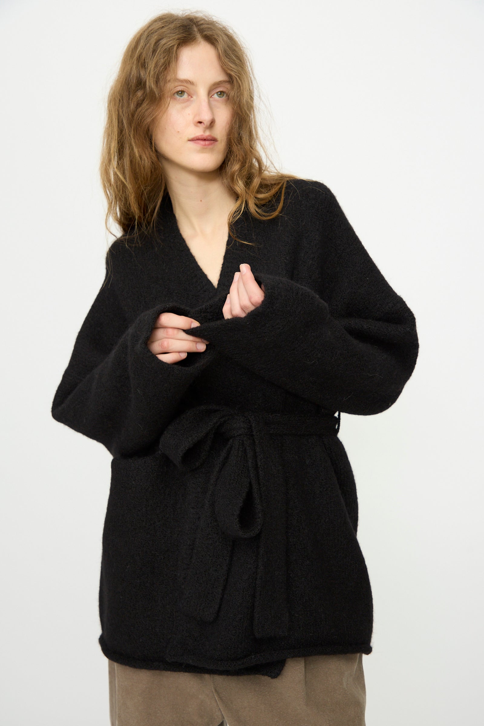 A person with long, light brown hair wears the Lauren Manoogian Coto Cardigan in Black and brown pants, standing against a plain white background.