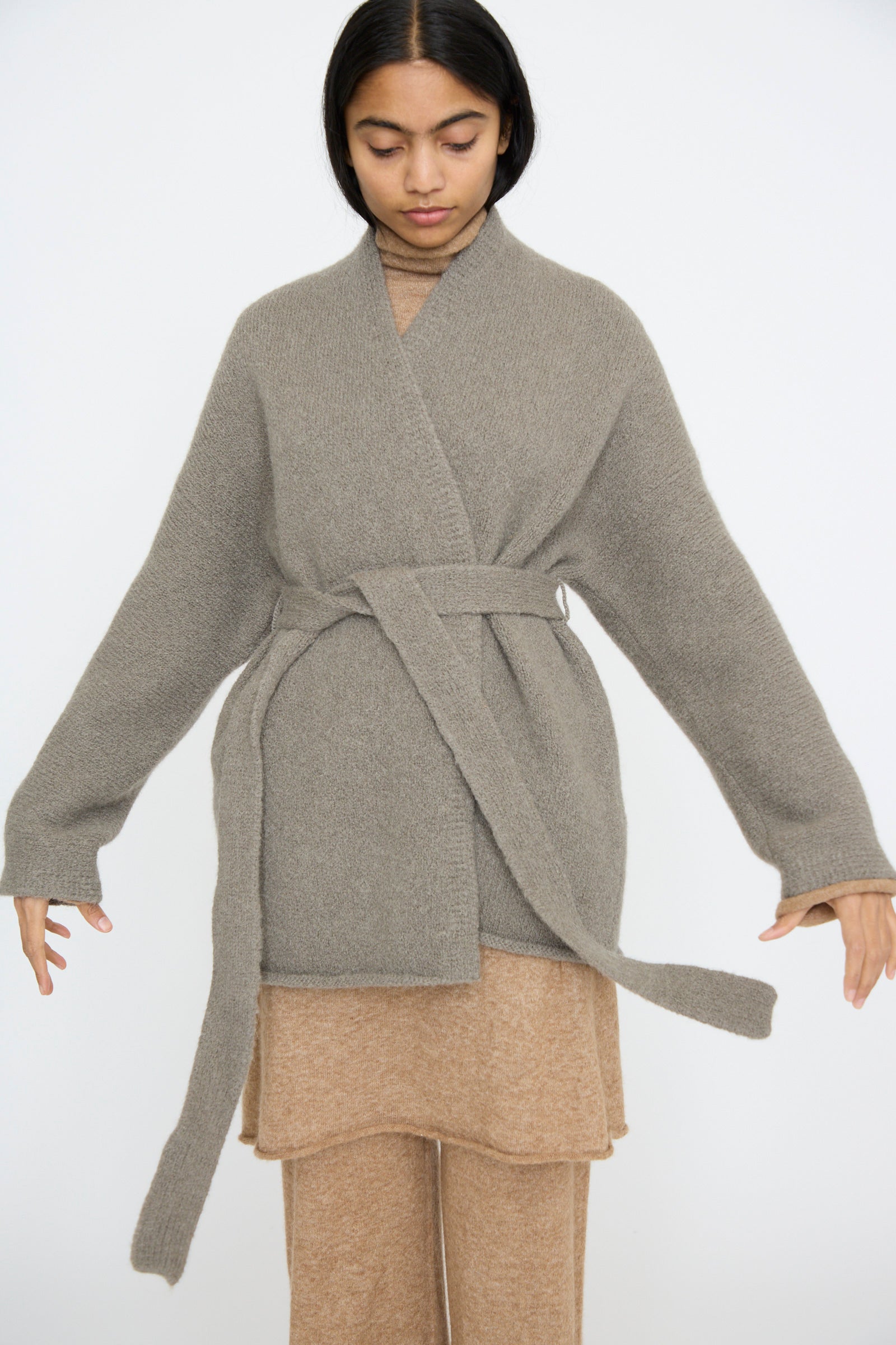 Against a plain backdrop, a person wears the Lauren Manoogian Coto Cardigan in Concrete, a gray hand-loomed alpaca wool piece with a belt, elegantly enhancing their beige outfit.