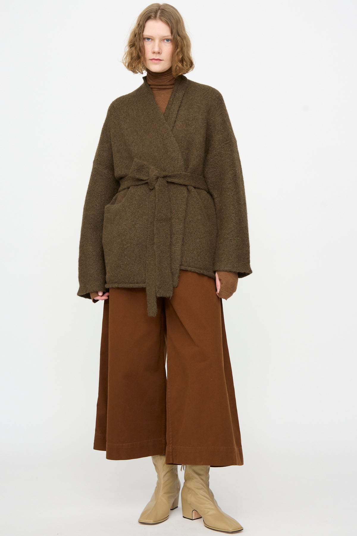 A person wearing the Lauren Manoogian Coto Cardigan in Olive—hand-loomed from alpaca in Peru—layered over a brown turtleneck, wide brown trousers, and beige ankle boots, standing against a plain white background.