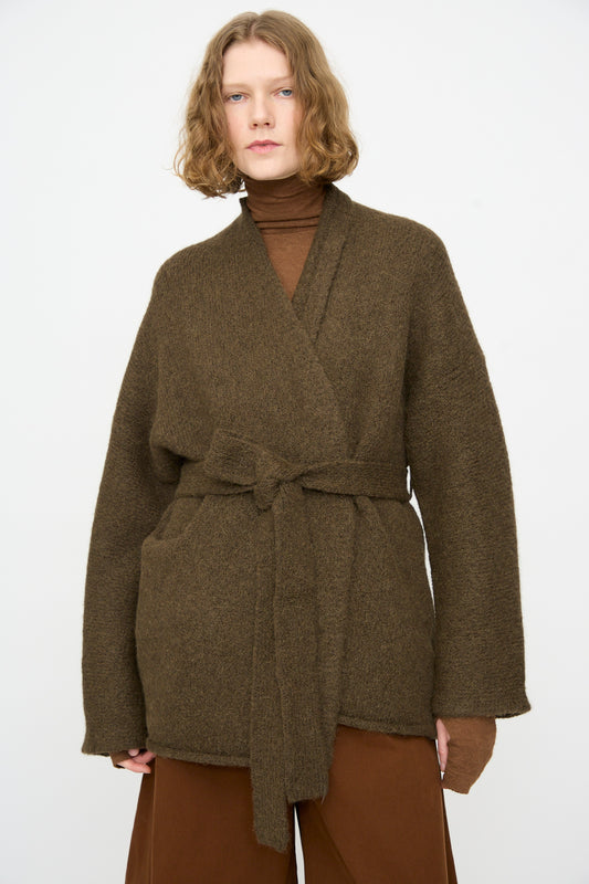 A person wearing a brown belted wrap coat over a brown turtleneck and brown pants, styled with the Lauren Manoogian Coto Cardigan in Olive—a hand-loomed alpaca piece made in Peru—stands against a plain white background.