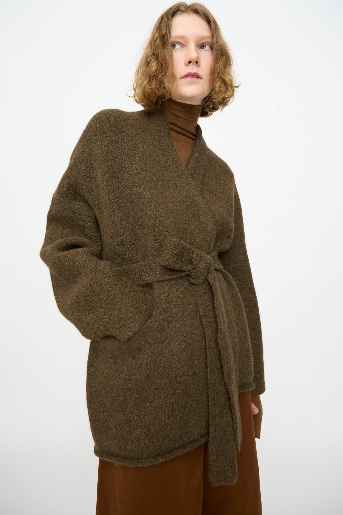 Person wearing the Lauren Manoogian Coto Cardigan in Olive, paired with a brown turtleneck and brown pants, standing against a plain white background.