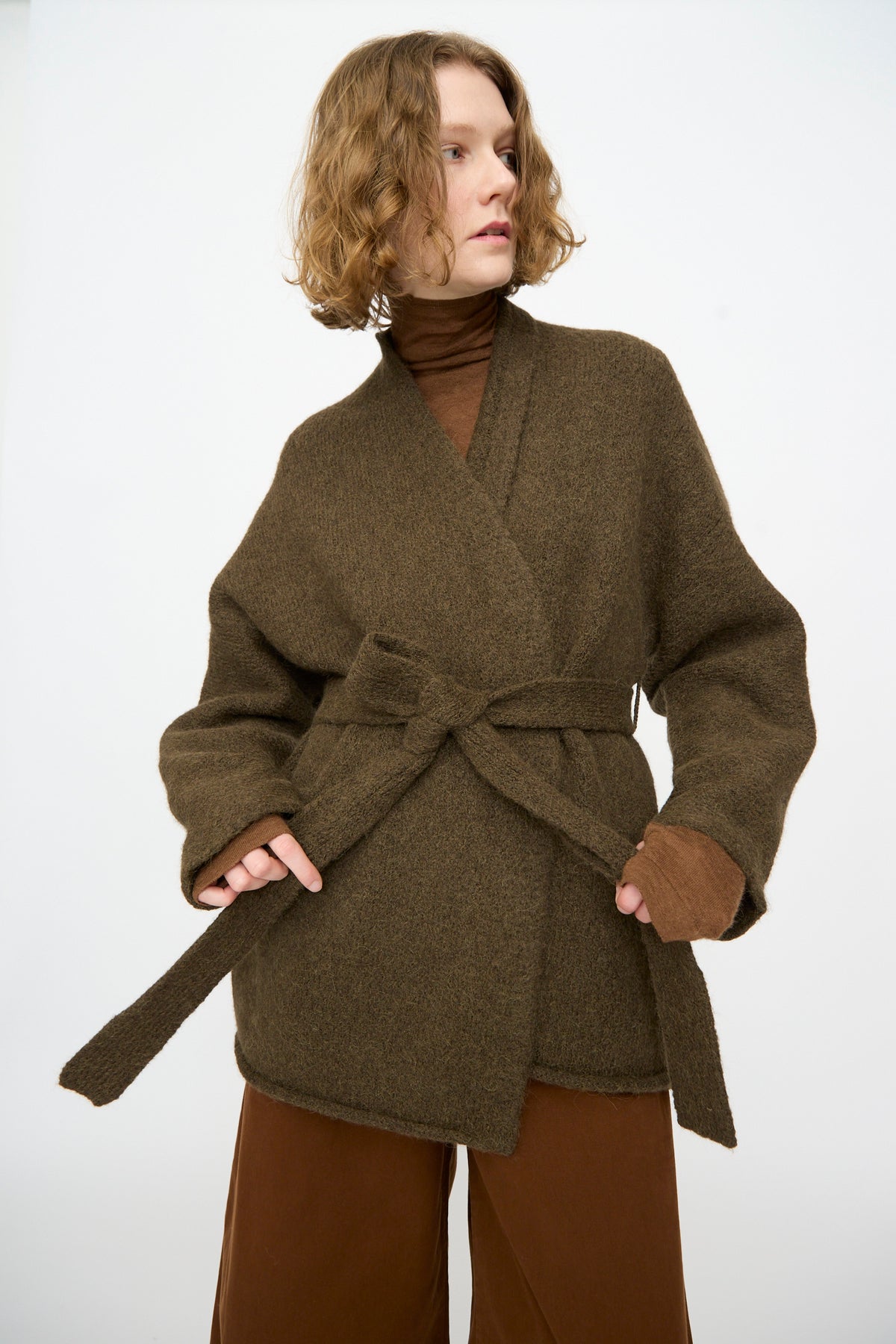 A person wearing Lauren Manoogian’s Coto Cardigan in Olive—belted and crafted from hand-loomed alpaca—over a brown turtleneck and wide-leg pants stands against a plain white background, adding warmth and texture to the look.