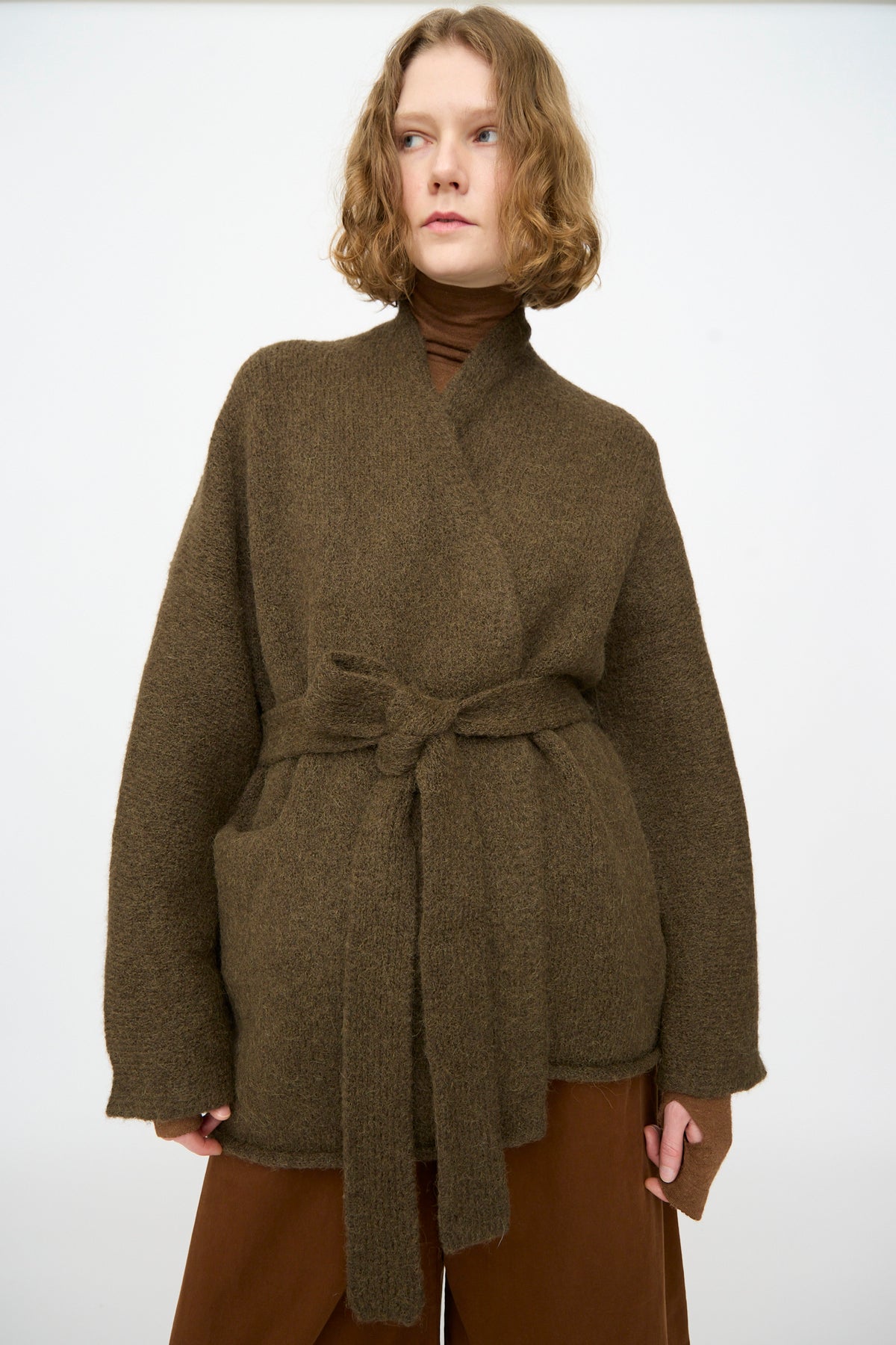 A person wearing the Lauren Manoogian Coto Cardigan in Olive, hand-loomed from alpaca in Peru, layered over a brown turtleneck and pants, stands against a plain white background.