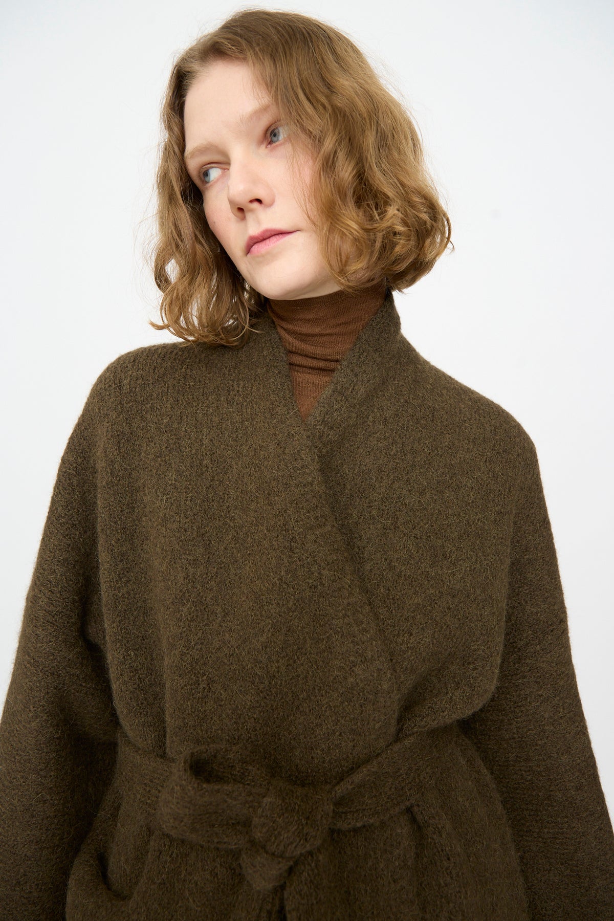 A woman with wavy light brown hair, dressed in a brown turtleneck and a belted dark brown wrap coat, layers her outfit with the Lauren Manoogian Coto Cardigan in Olive—a hand-loomed alpaca piece made in Peru—while looking slightly to the side against a plain background.