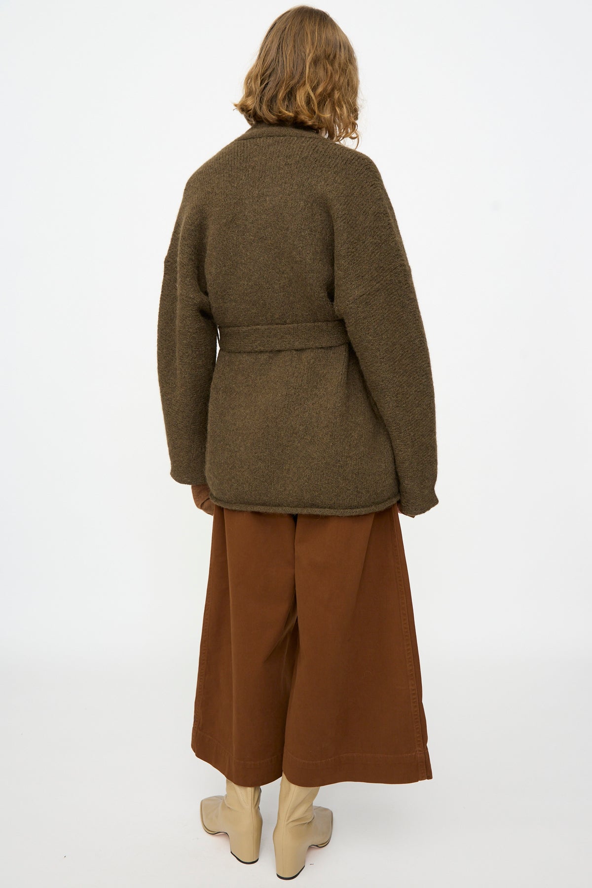 A person stands with their back to the camera, wearing the Lauren Manoogian Coto Cardigan in Olive—a belted, hand-loomed alpaca piece crafted in Peru—styled with wide brown pants and cream boots, set against a plain white background.