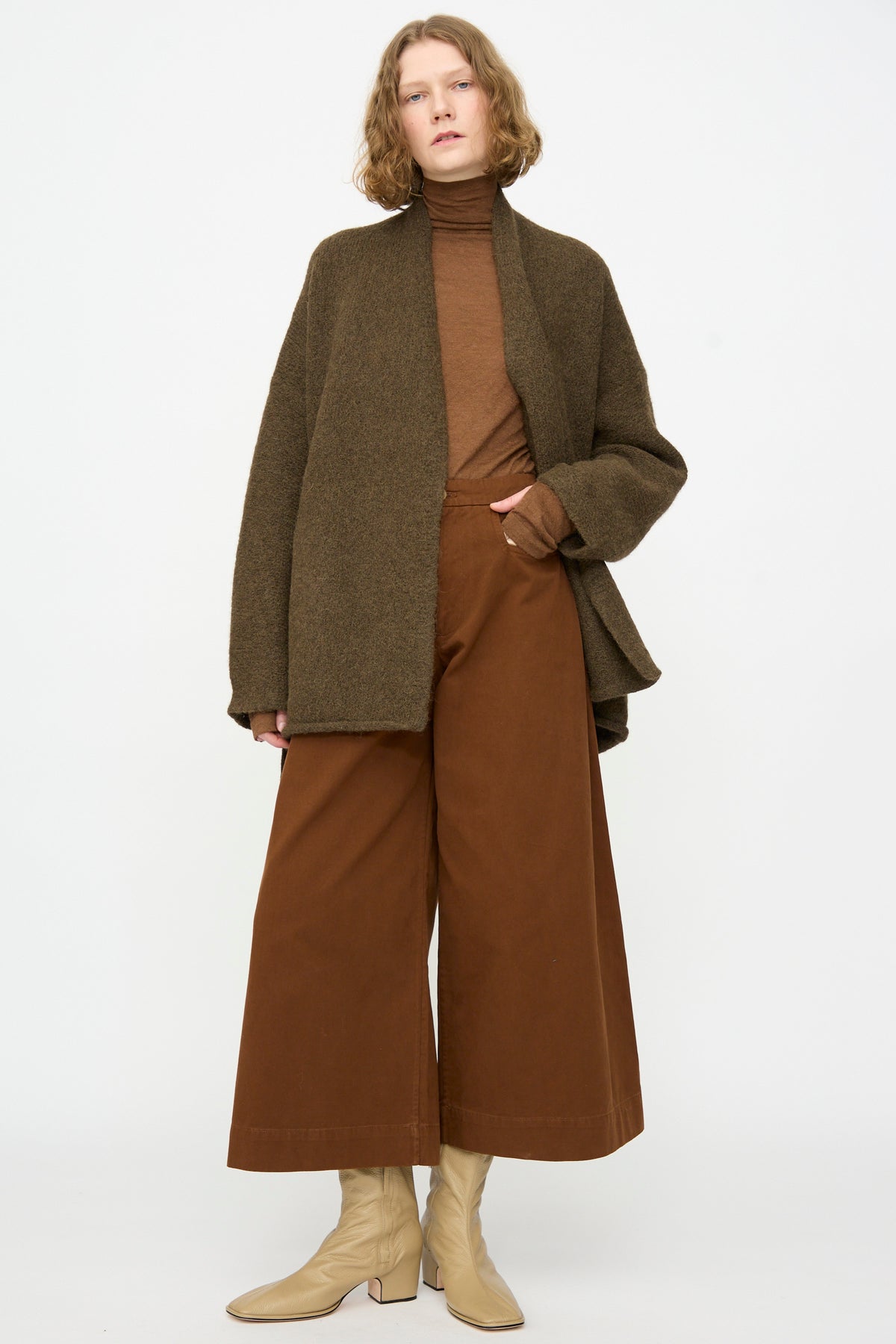 A person wearing a brown turtleneck, the Lauren Manoogian Coto Cardigan in Olive, wide-leg brown pants, and beige ankle boots stands against a plain white background.