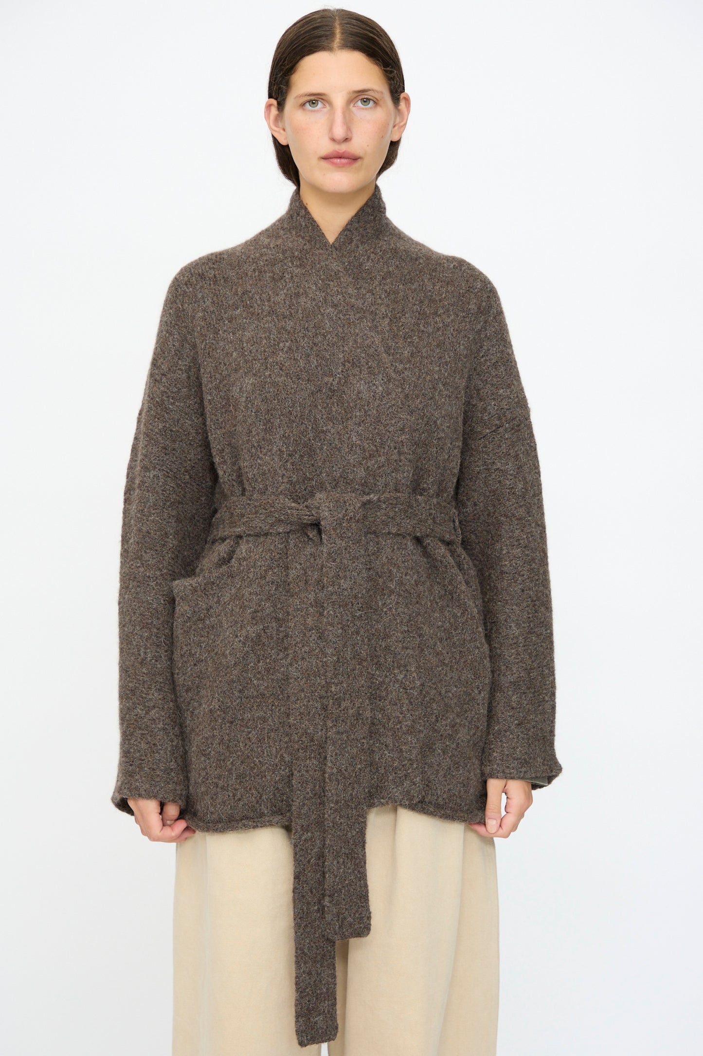 A woman faces forward against a white background, wearing the Lauren Manoogian Coto Cardigan in Patina—a hand-loomed brown belted alpaca cardigan with a relaxed fit—paired with cream-colored pants.