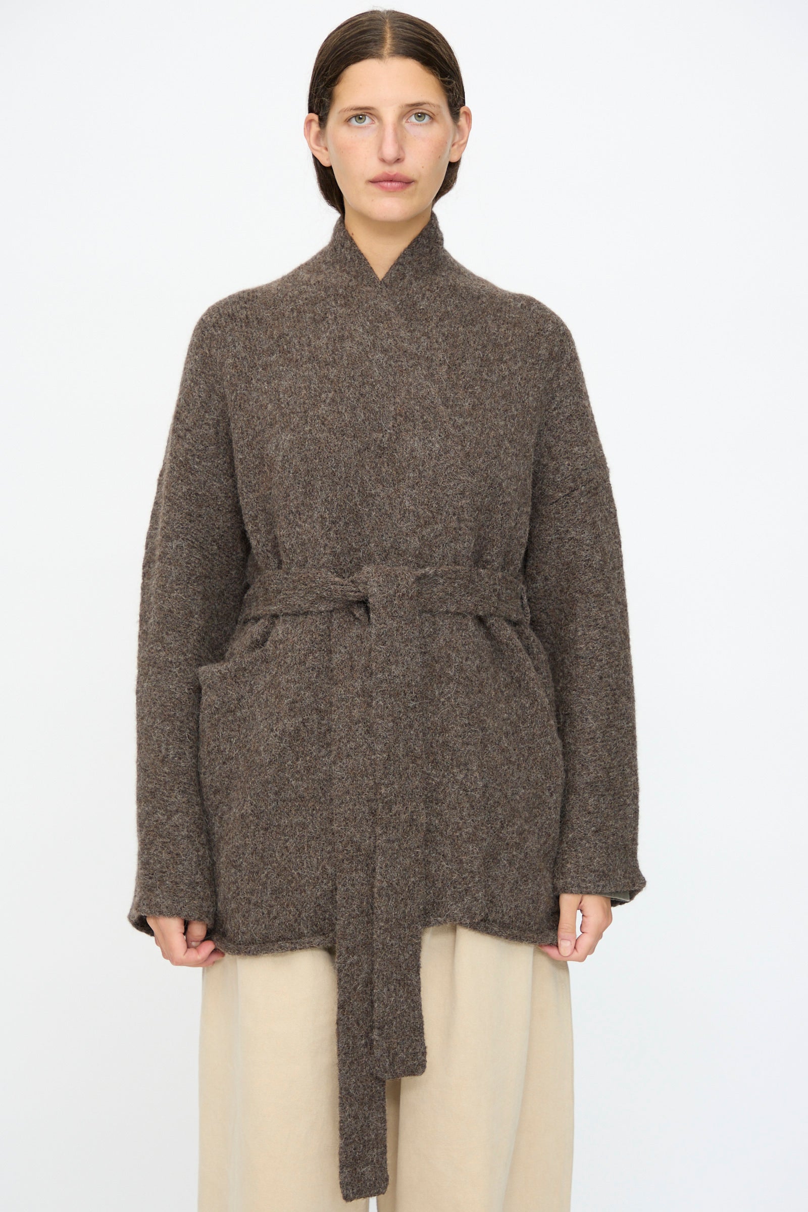A woman faces forward against a white background, wearing the Lauren Manoogian Coto Cardigan in Patina—a hand-loomed brown belted alpaca cardigan with a relaxed fit—paired with cream-colored pants.