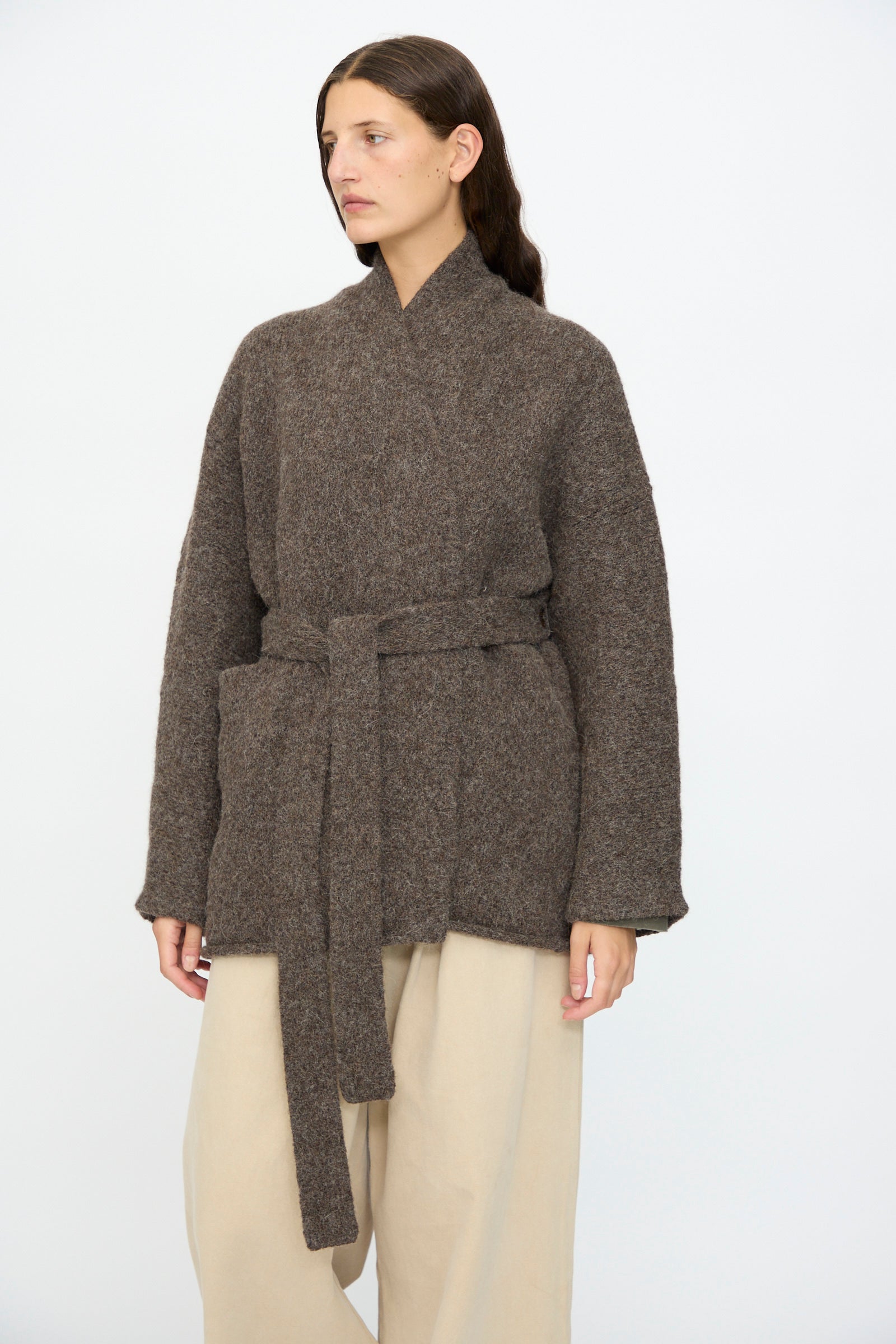A woman stands against a plain white background wearing Lauren Manoogian’s Coto Cardigan in Patina, a hand-loomed alpaca piece with a relaxed fit, paired with beige wide-leg pants.