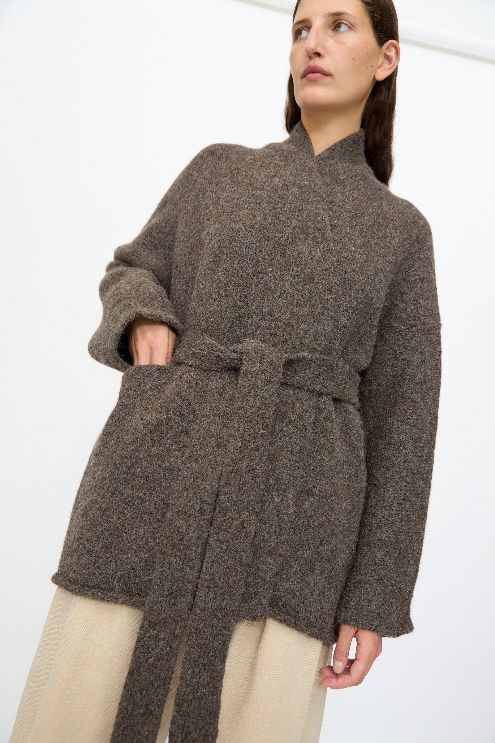 A person wears the Lauren Manoogian Coto Cardigan in Patina, a relaxed fit hand-loomed brown wool belted cardigan, over beige pants, standing against a plain white background.