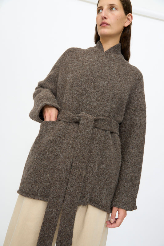 A person wears the Lauren Manoogian Coto Cardigan in Patina, a relaxed fit hand-loomed brown wool belted cardigan, over beige pants, standing against a plain white background.