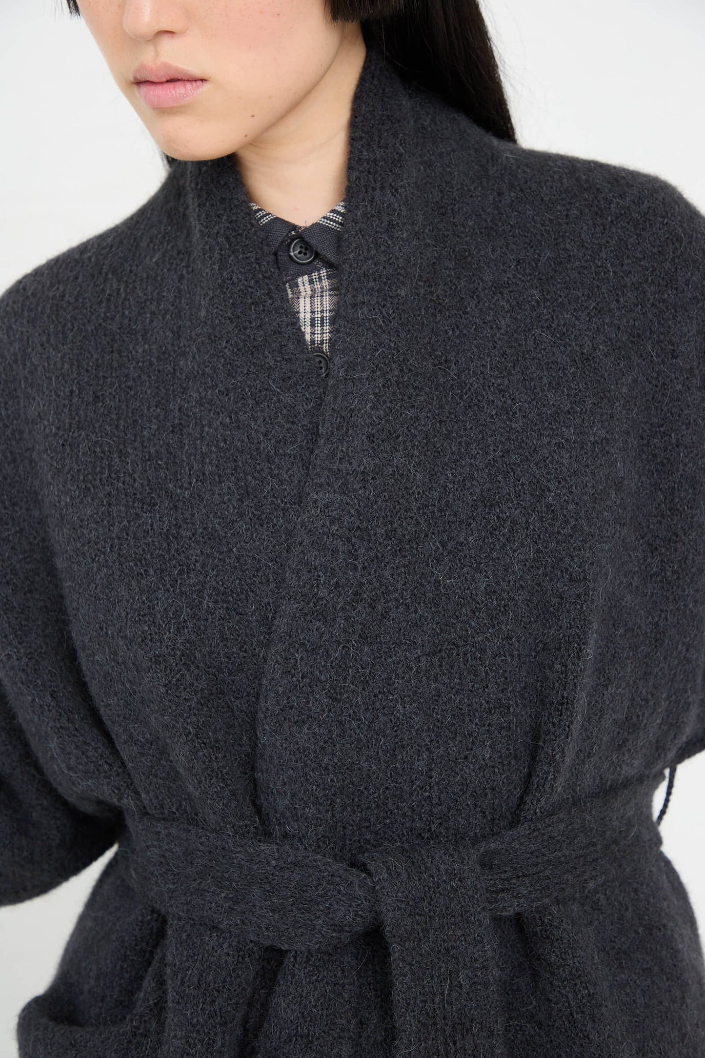 A person wearing a dark gray, textured wool coat with a belted waist over a collared shirt, reminiscent of the refined style of the Coto Cardigan in Ink by Lauren Manoogian.