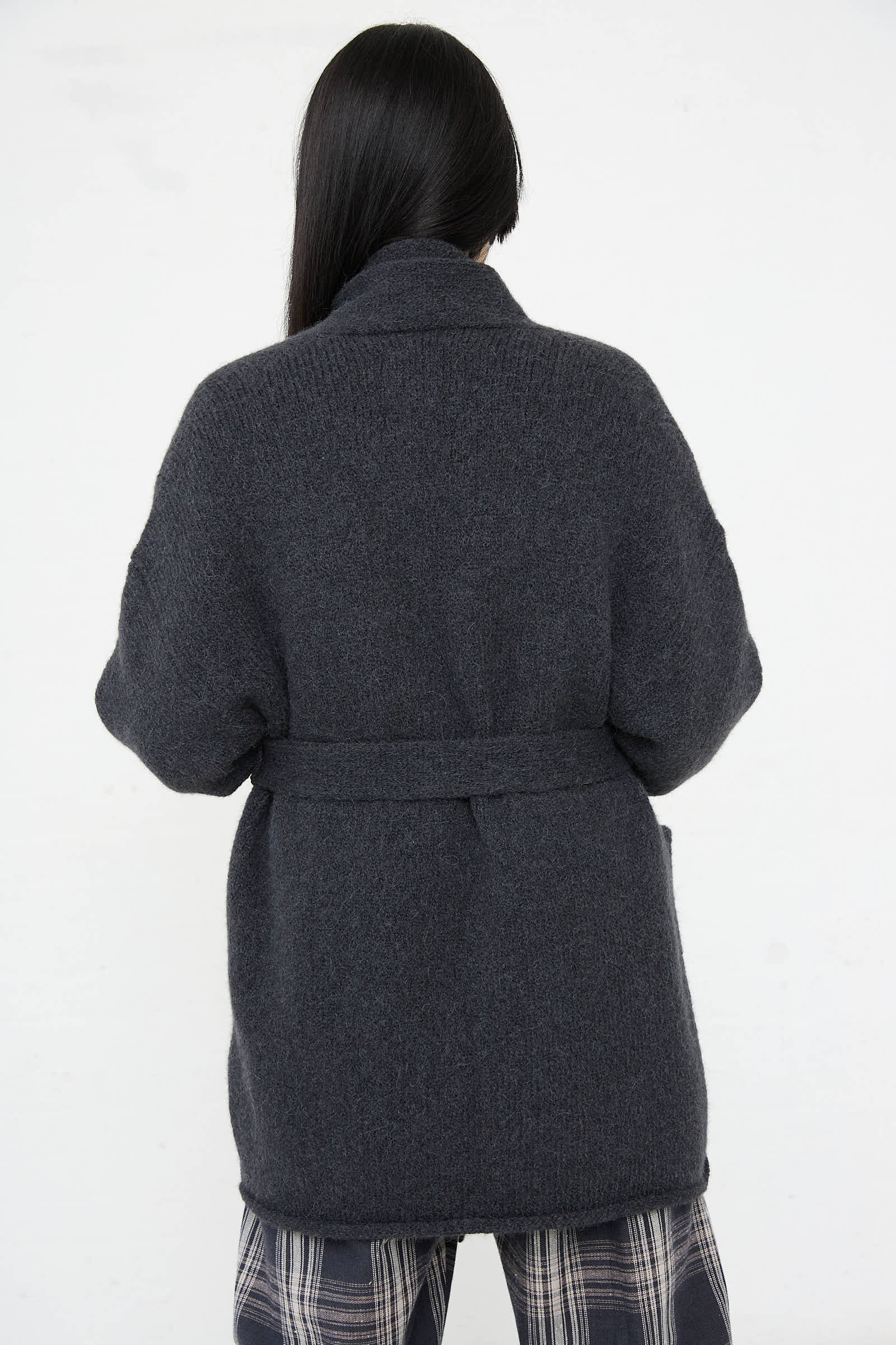 A person with long, dark hair wearing a belted, dark gray wool coat and plaid pants, photographed from behind against a plain white background, exudes a timeless elegance reminiscent of the Lauren Manoogian Coto Cardigan in Ink.