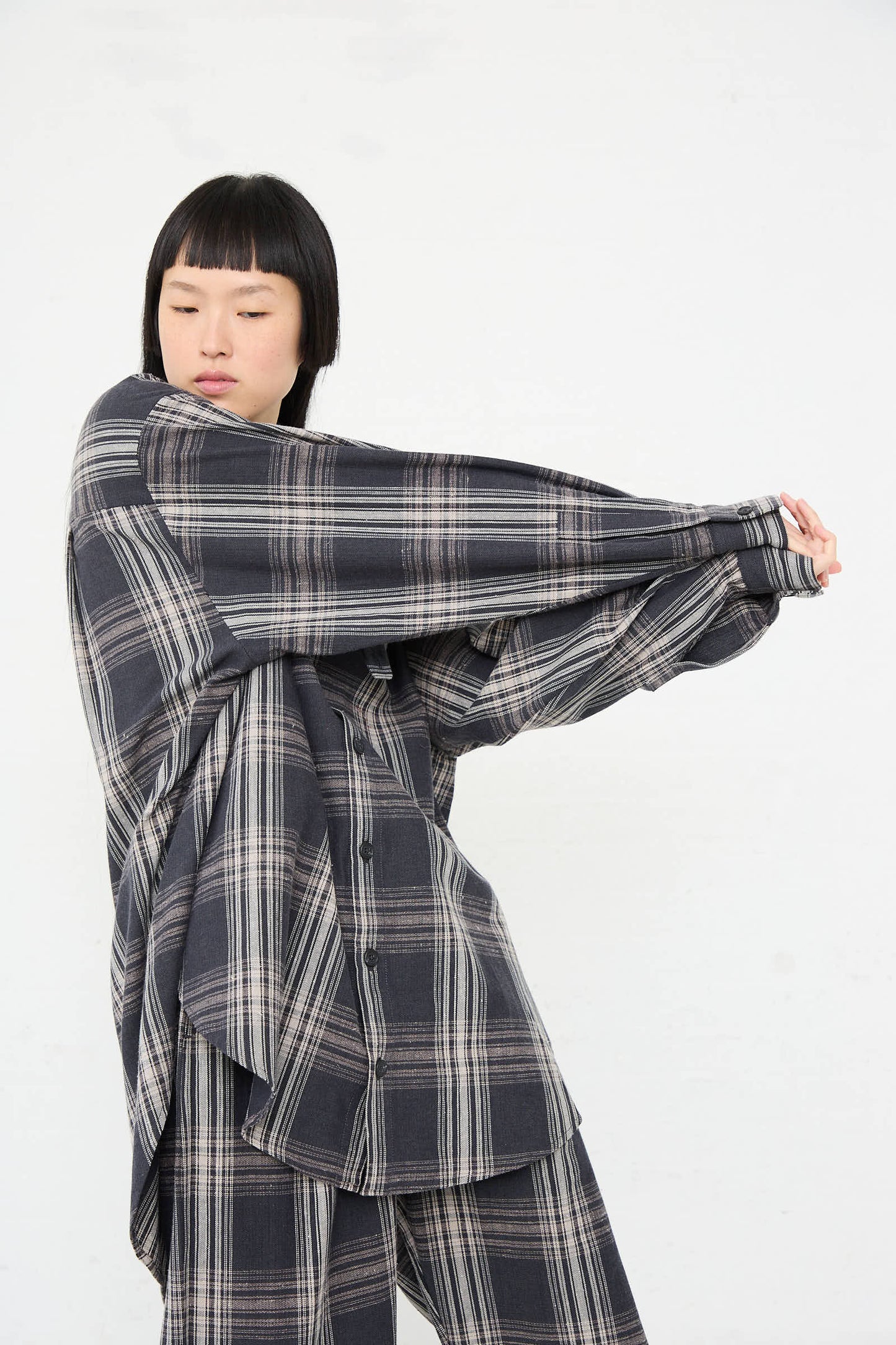 A person wearing the Lauren Manoogian Cotton Gaiter Shirt in Plaid, an oversized black and white soft cotton shirt, stretches their right arm across their chest against a white background.