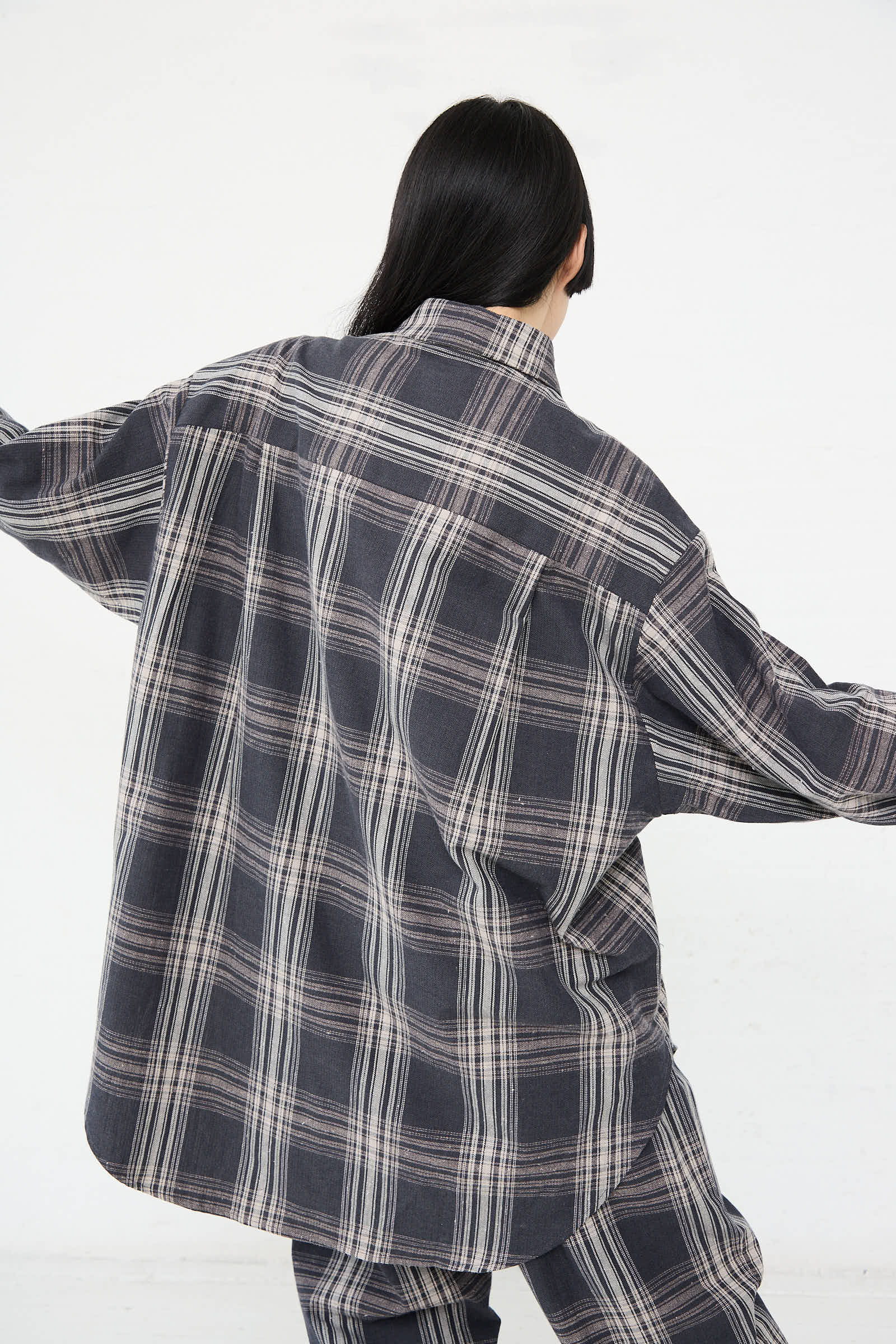 A person with long dark hair is facing away from the camera, wearing an oversized Lauren Manoogian Cotton Gaiter Shirt in Plaid with matching pants. The background is plain white.