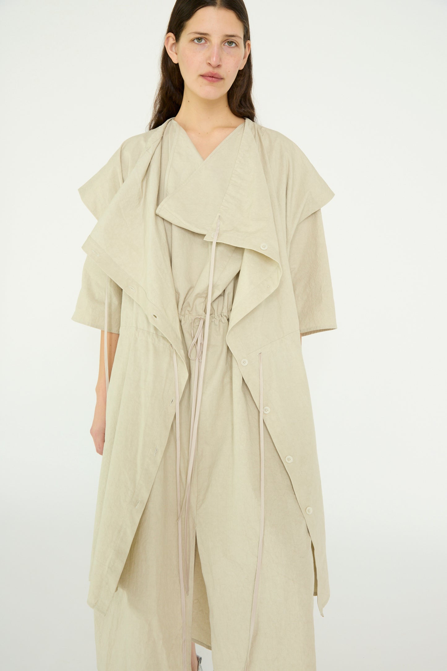 A person wearing Lauren Manoogian's Cowl Gilet in Plaster, featuring a beige layered design, asymmetrical cut, drawstring waist, and oversized fit, stands against a plain background.