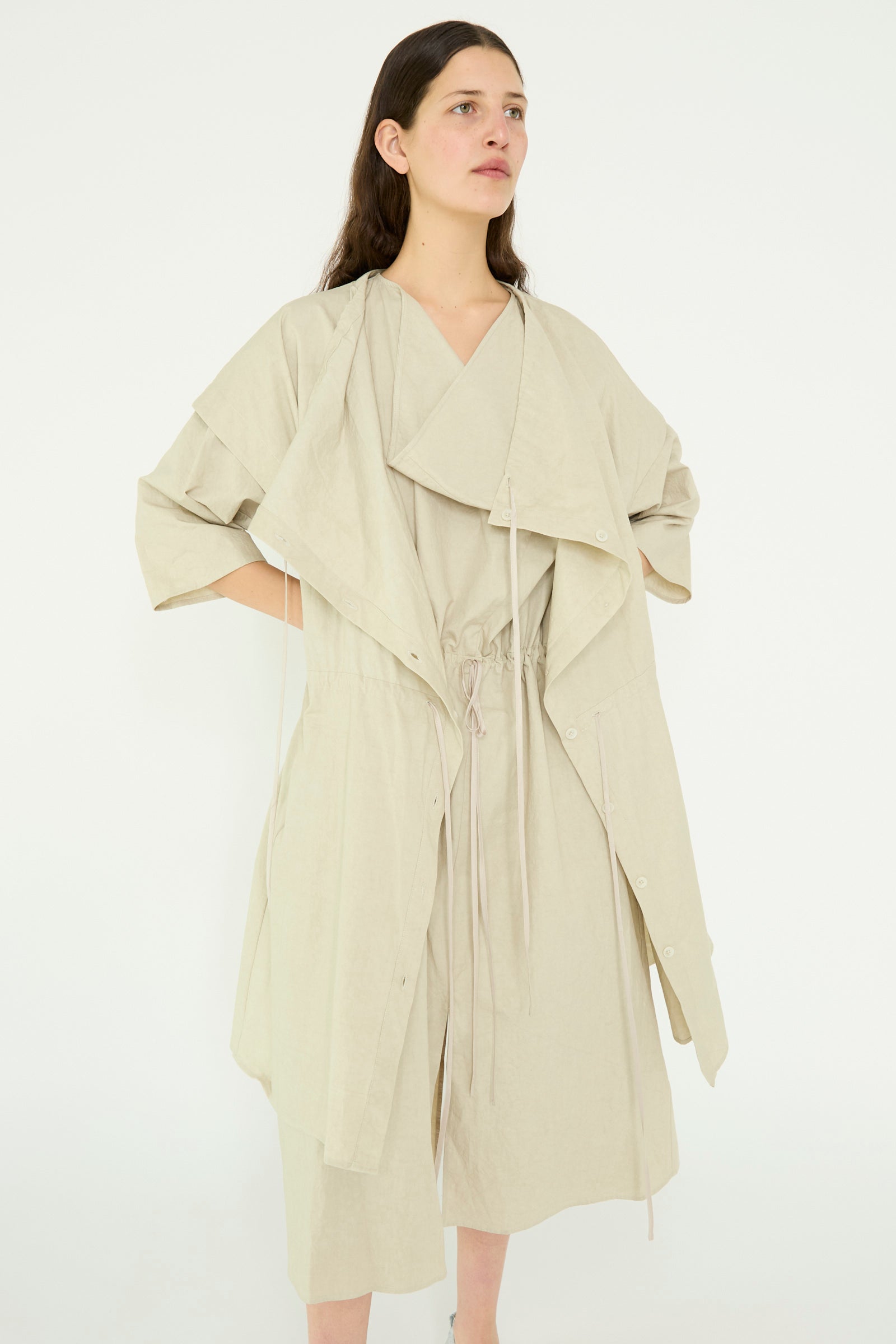 A person wears a layered light beige dress with ruffled sleeves and drawstring details, paired with the Lauren Manoogian Cowl Gilet in Plaster, standing against a plain background.