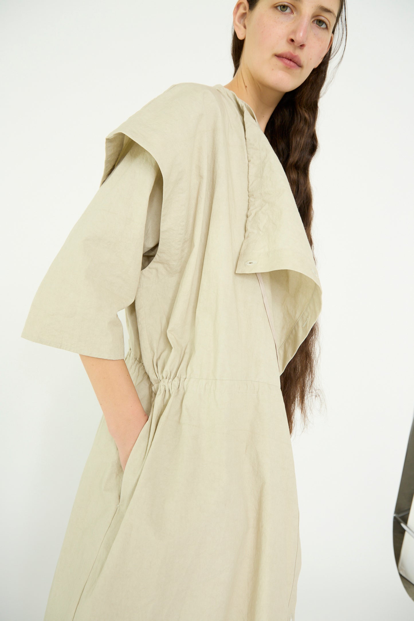 A person, clad in the Lauren Manoogian Cowl Gilet in Plaster, poses against a plain background with a hand casually tucked into one of its pockets, showcasing its loose and oversized fit.